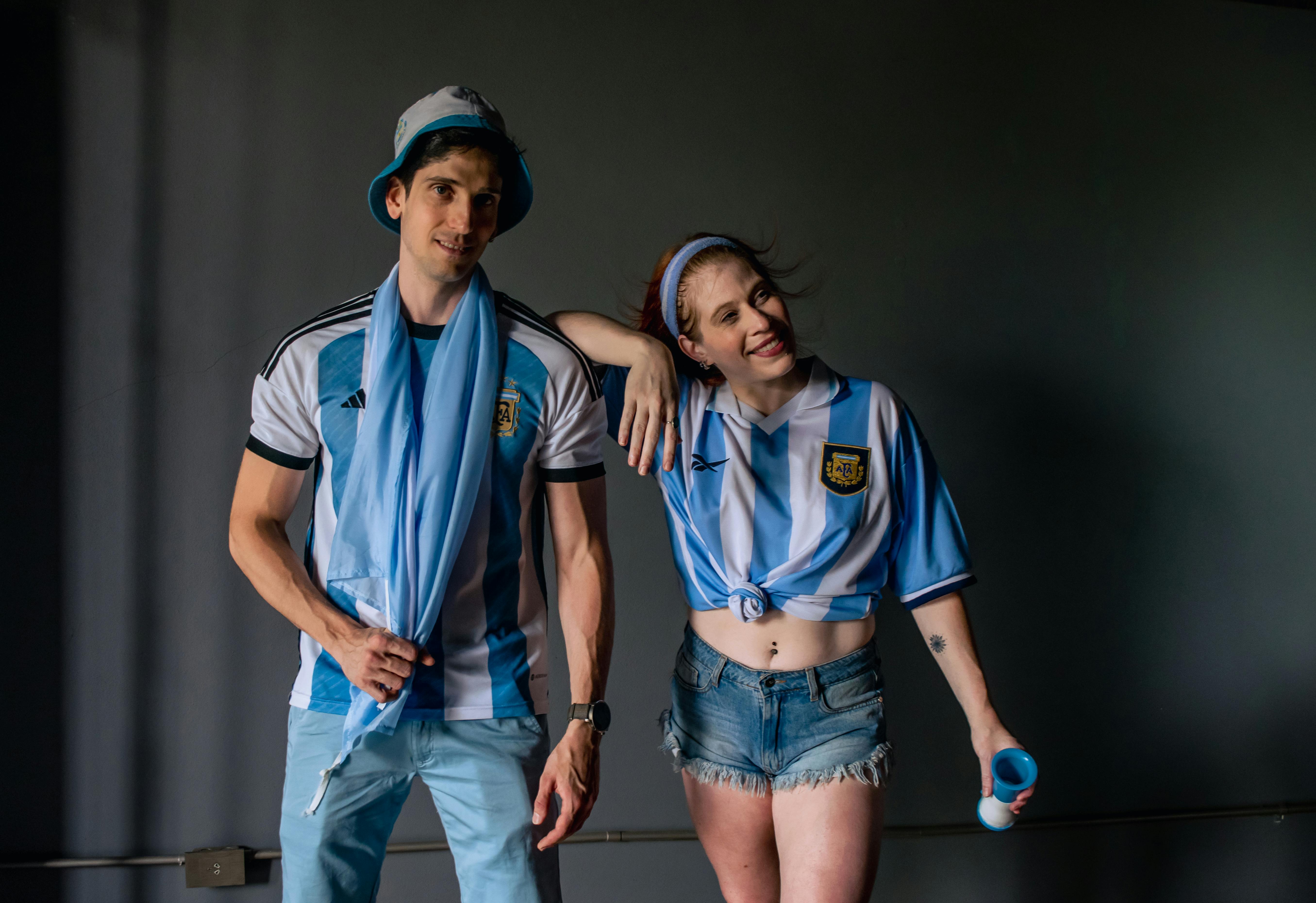 Two young fans in Argentina soccer apparel pose with enthusiasm indoors.