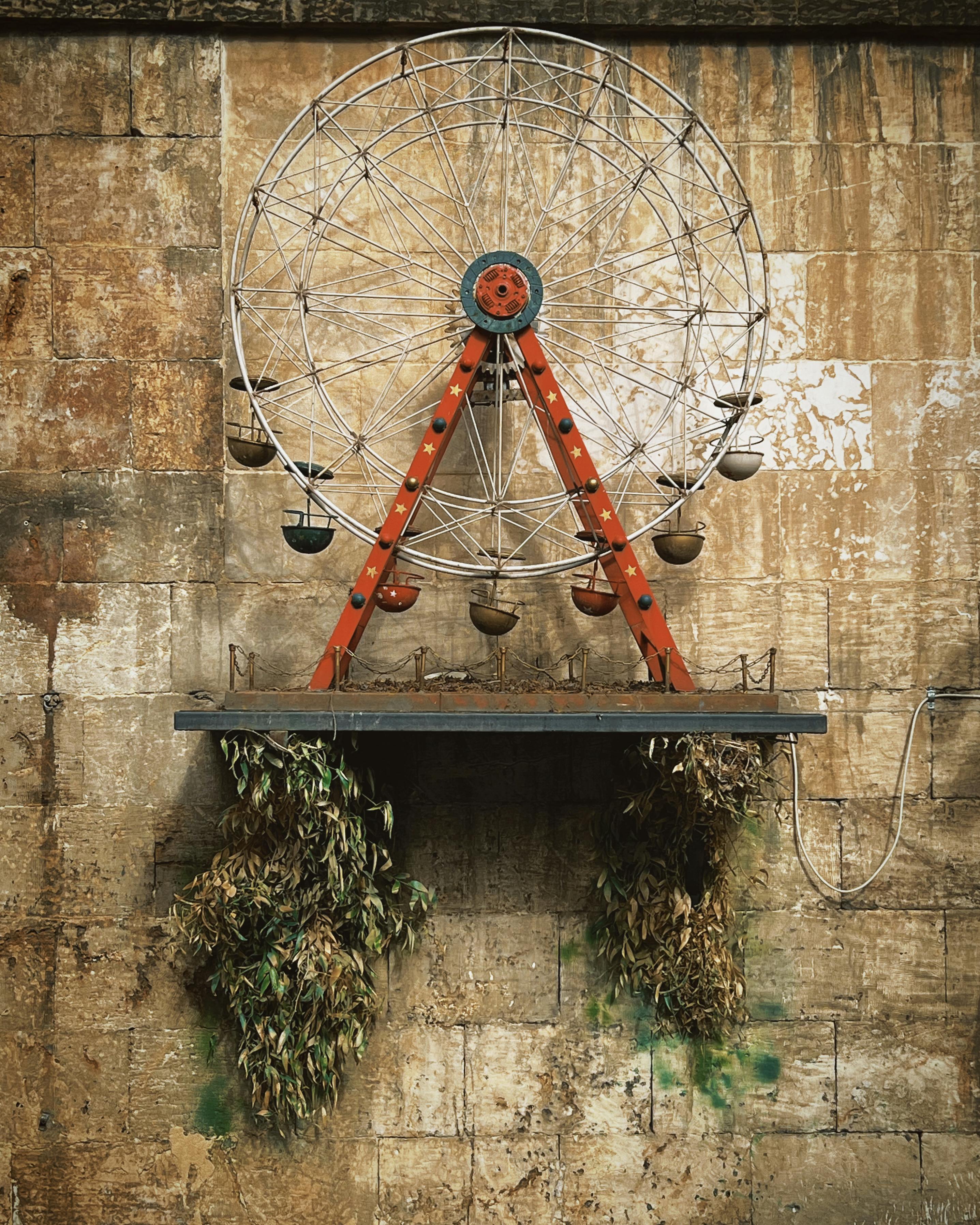 бесплатная Небольшое декоративное колесо обозрения, установленное на кирпичной стене в деревенском стиле, с растениями внизу, создающее винтажную атмосферу. Стоковое фото