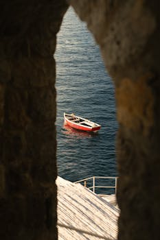 Red boat floating by the coast viewed through an ancient stone archway.
