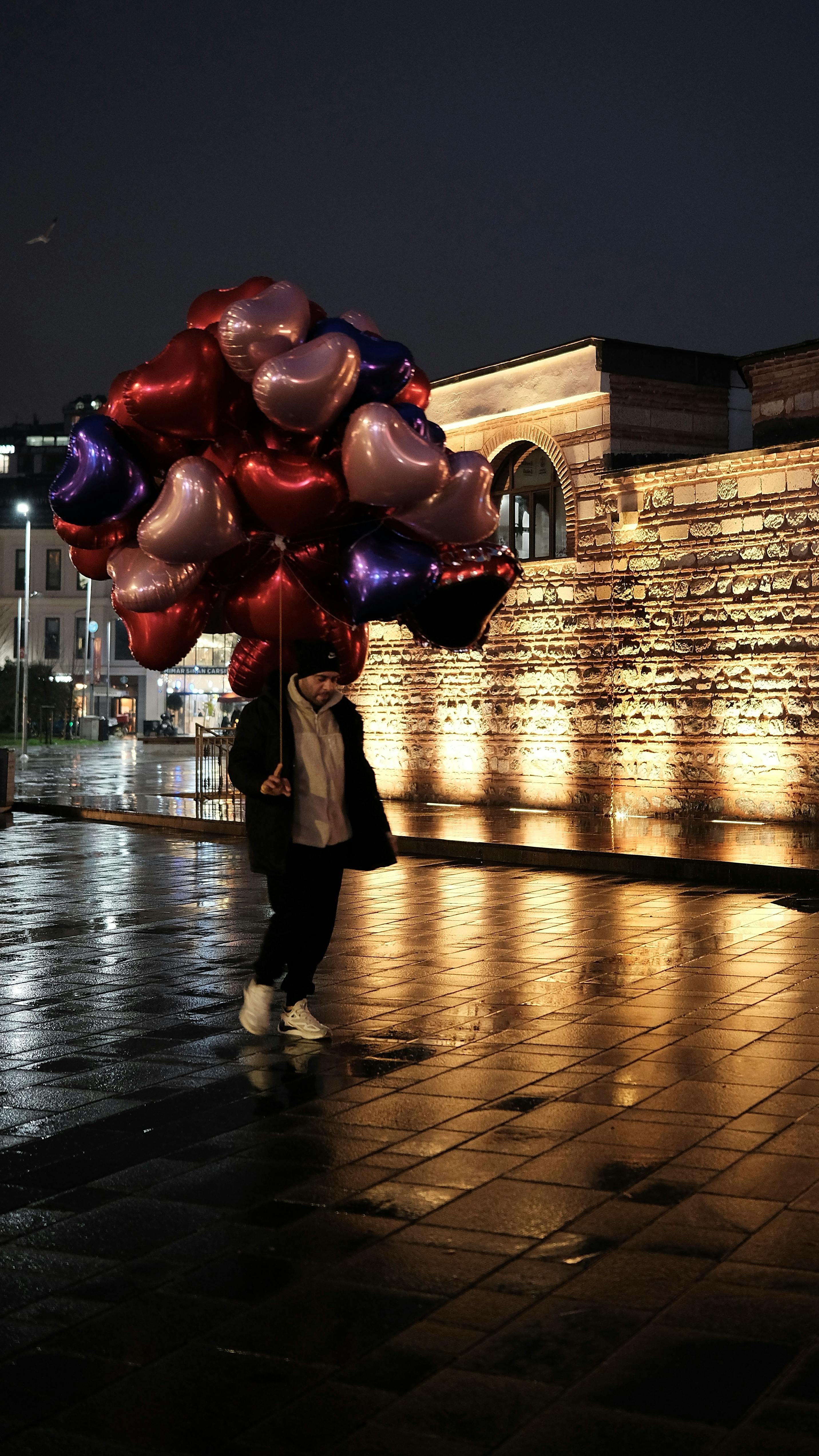 бесплатная Ночью в Стамбуле человек несет воздушные шары в форме сердца по мокрой улице. Стоковое фото