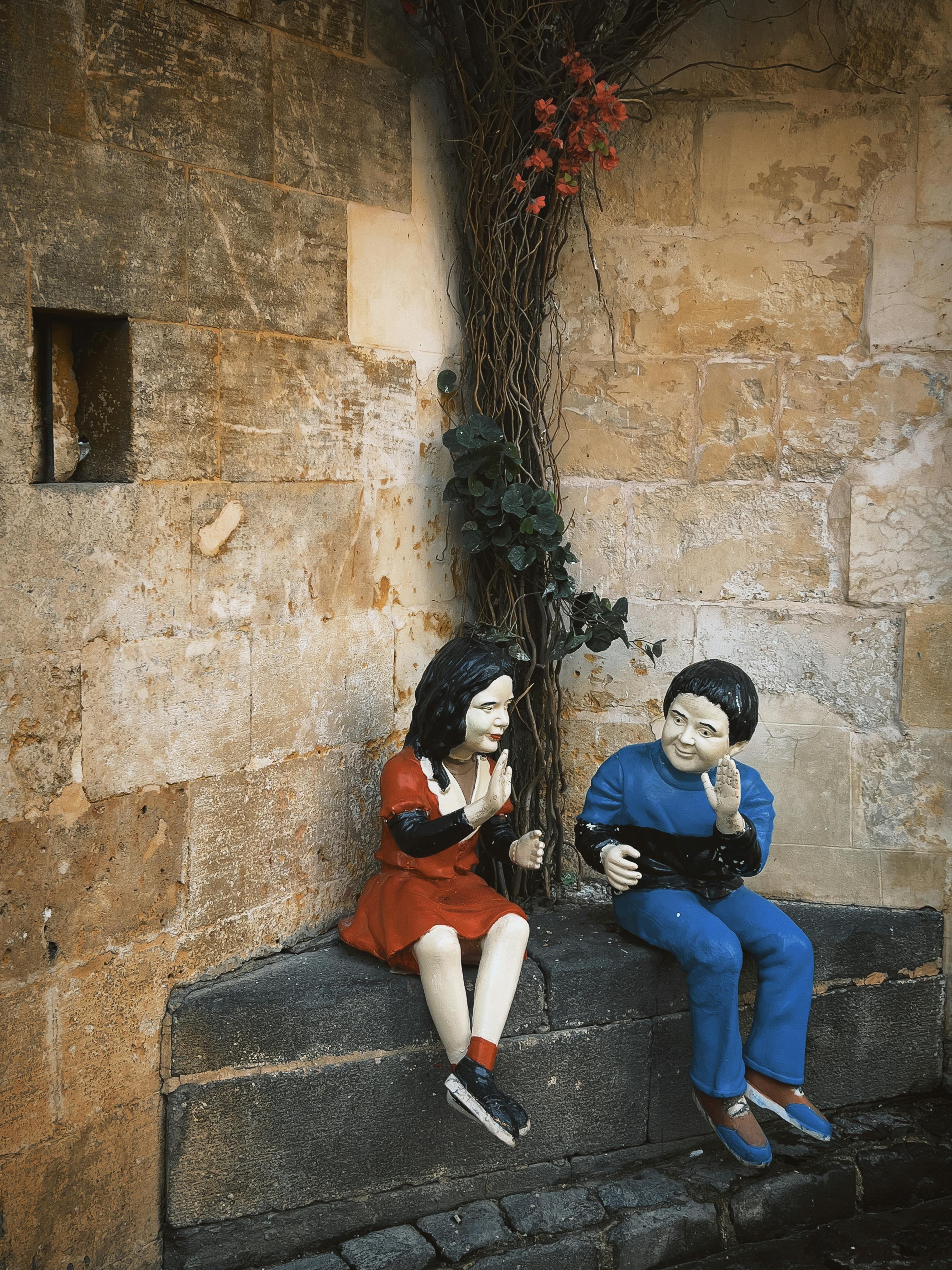 ingyenes Két gyermek élénk színű szobra, akik egy párkányon ülnek egy indákkal szegélyezett kőfalnak támasztva. Stockfotó