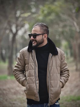 Fashionable man with sunglasses in a quilted jacket enjoying a walk in a park setting.