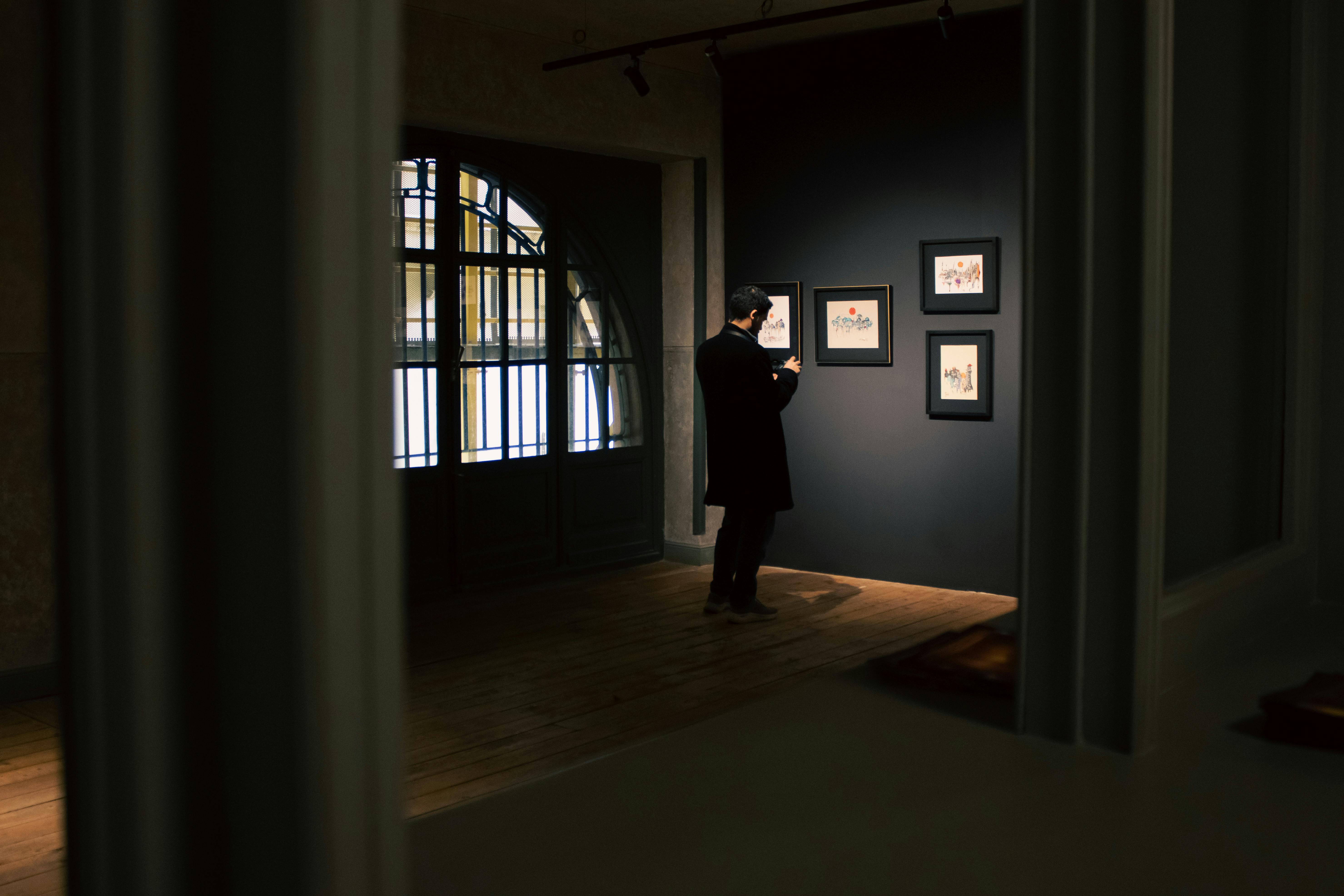 A man in a gallery analyzes framed art in dim lighting, creating a sophisticated atmosphere.