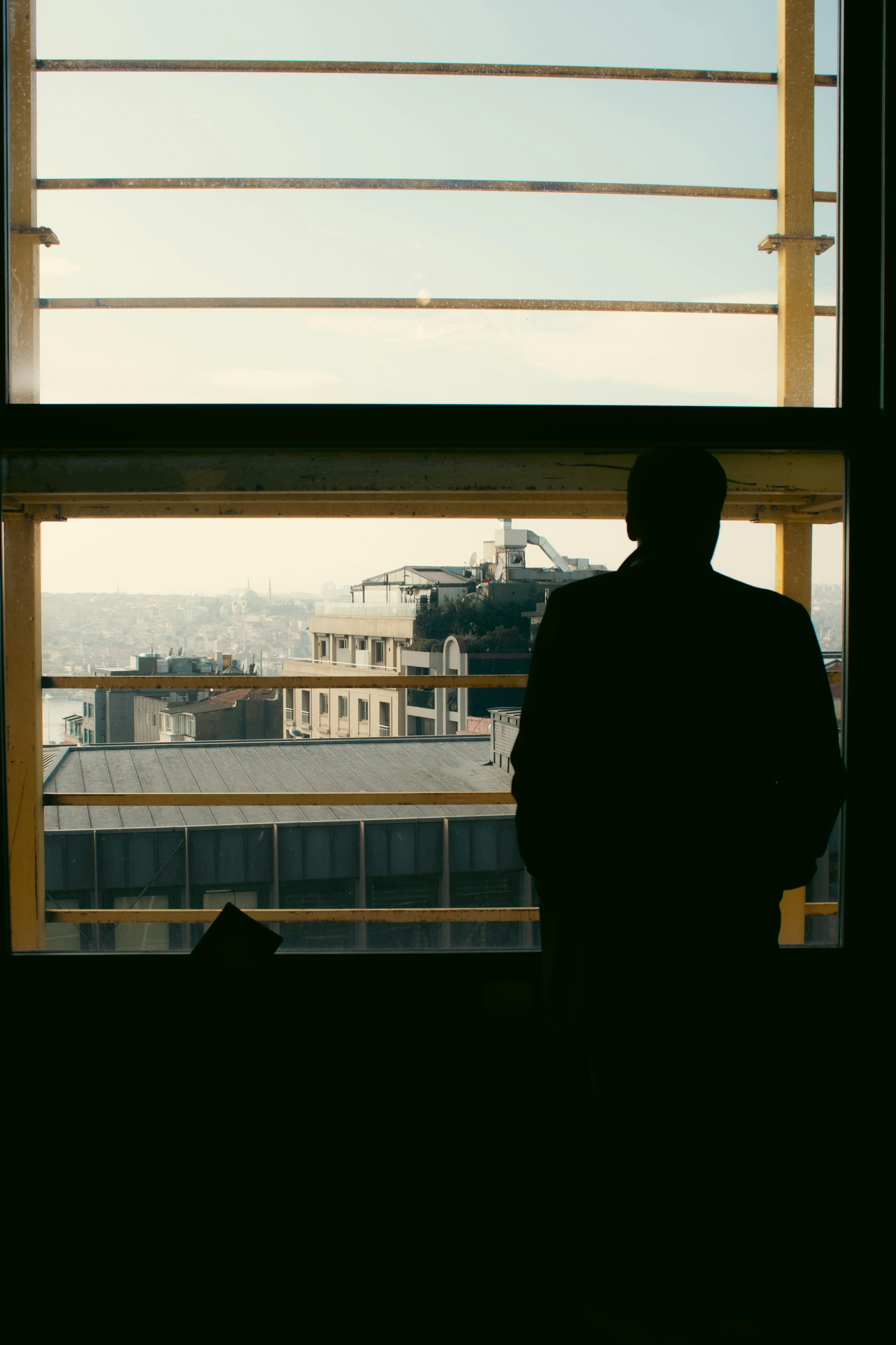Δωρεάν Σιλουέτα ενός άνδρα που στέκεται δίπλα σε ένα παράθυρο, επιβλέποντας τα κτίρια της πόλης στο φως της ημέρας. Φωτογραφία από στοκ φωτογραφιών