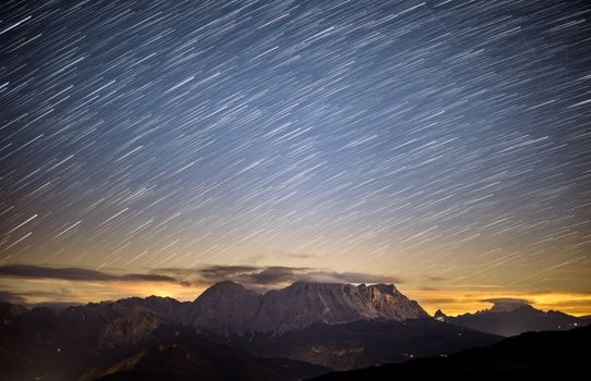 Dramatic long exposure of star trails above the Dolomite mountains at sunset, showcasing a stunning natural landscape.