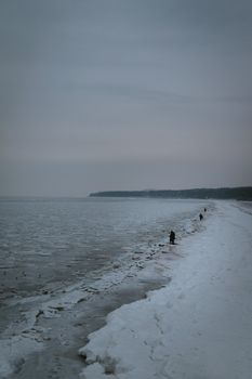 Distant figures walk along a snowy, frozen beach under an overcast evening sky.