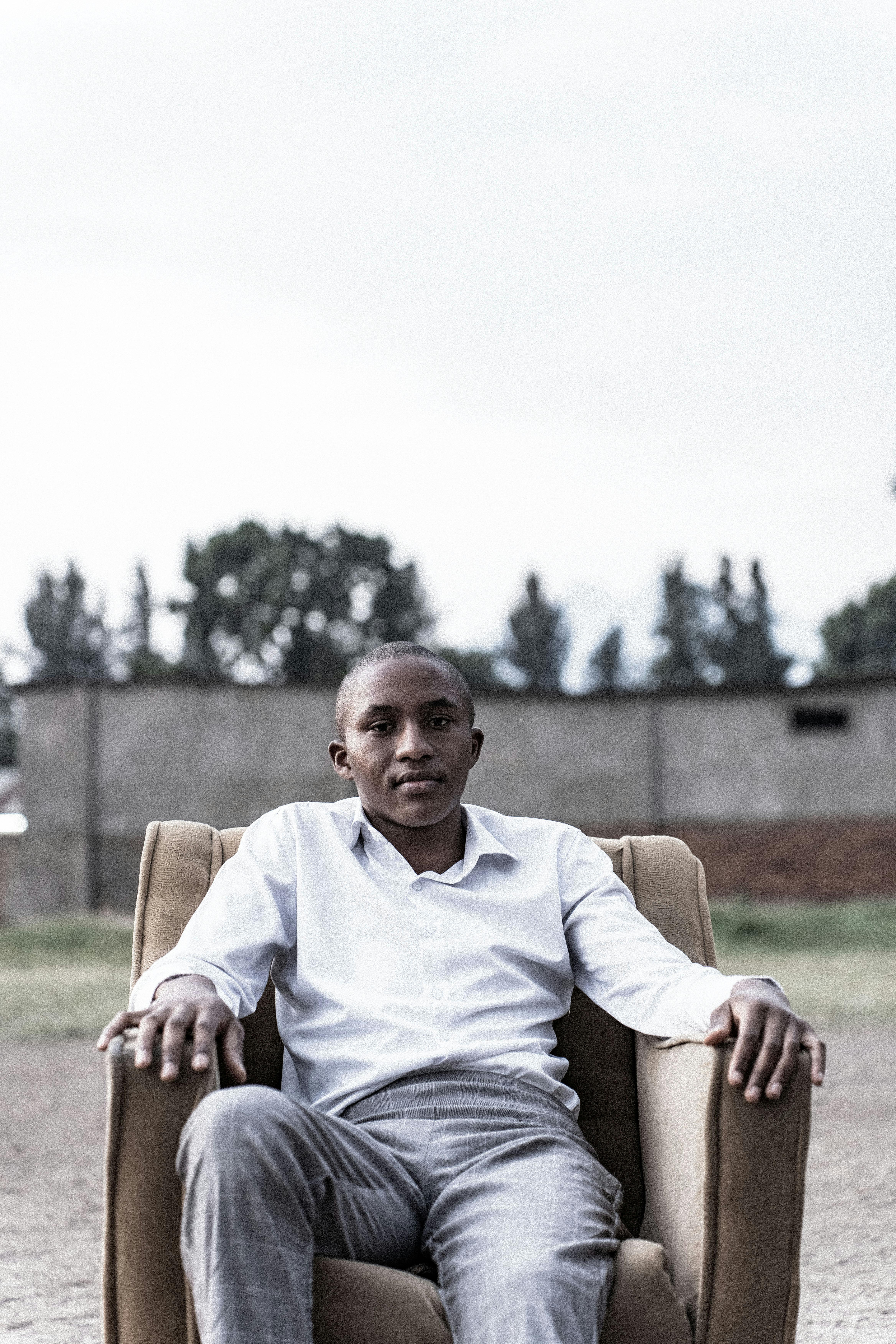 Zadarmo Mladý muž sedí vonku na stoličke, oblečený v ležérnej bielej košeli a sivých nohaviciach. Fotka z fotobanky