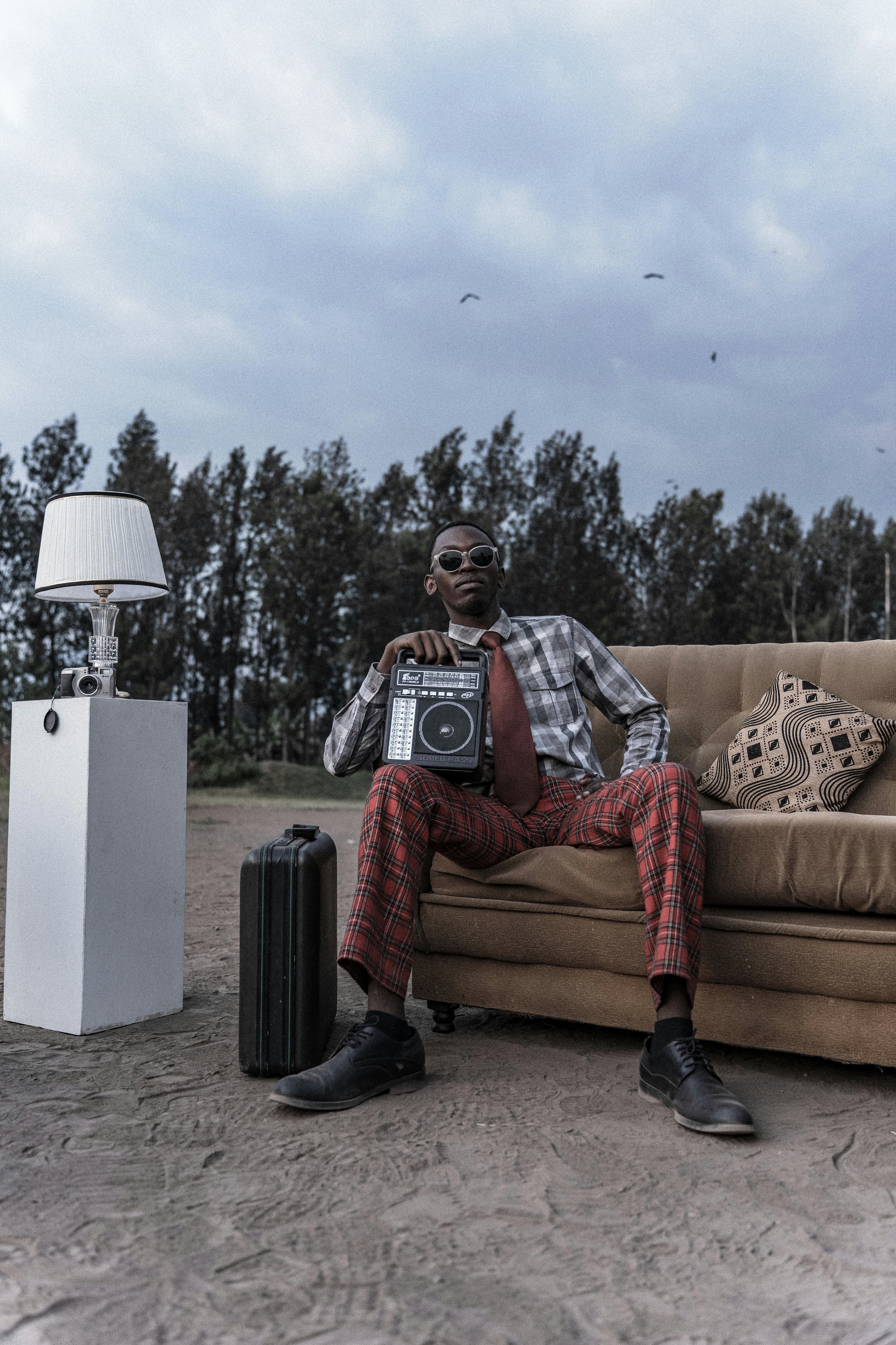 Miễn phí Một người đàn ông sành điệu ngồi ngoài trời trên ghế dài, bên cạnh là chiếc radio cổ, vali và đèn, thể hiện phong cách retro và tinh thần du lịch. Ảnh lưu trữ