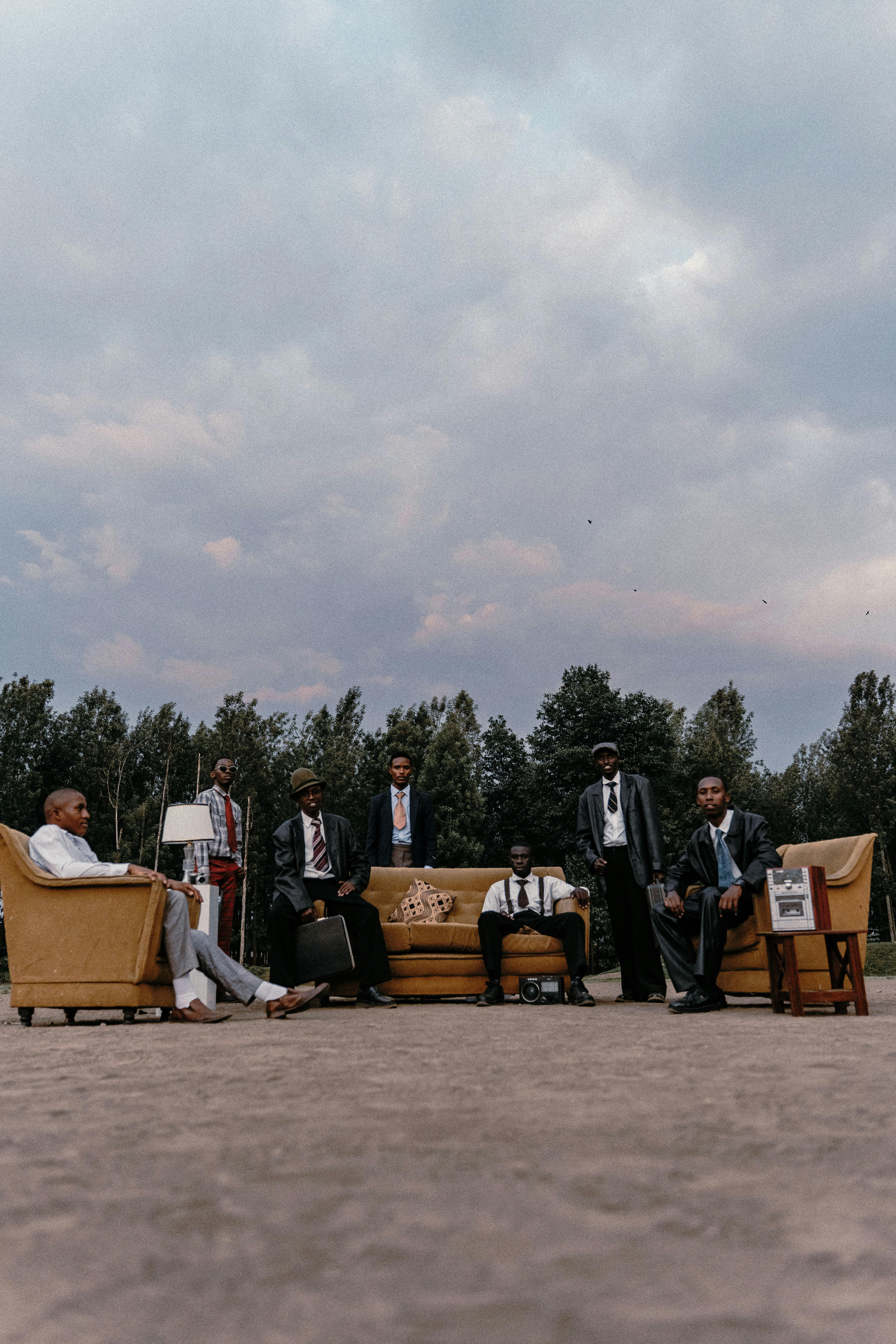 Free A stylish group of men in suits seated outdoors with vintage furniture under a cloudy sky. Stock Photo