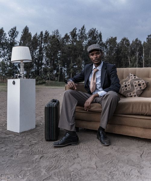 Free A stylish man sitting on a couch outdoors, flanked by a suitcase and a lamp in a rustic setting. Stock Photo