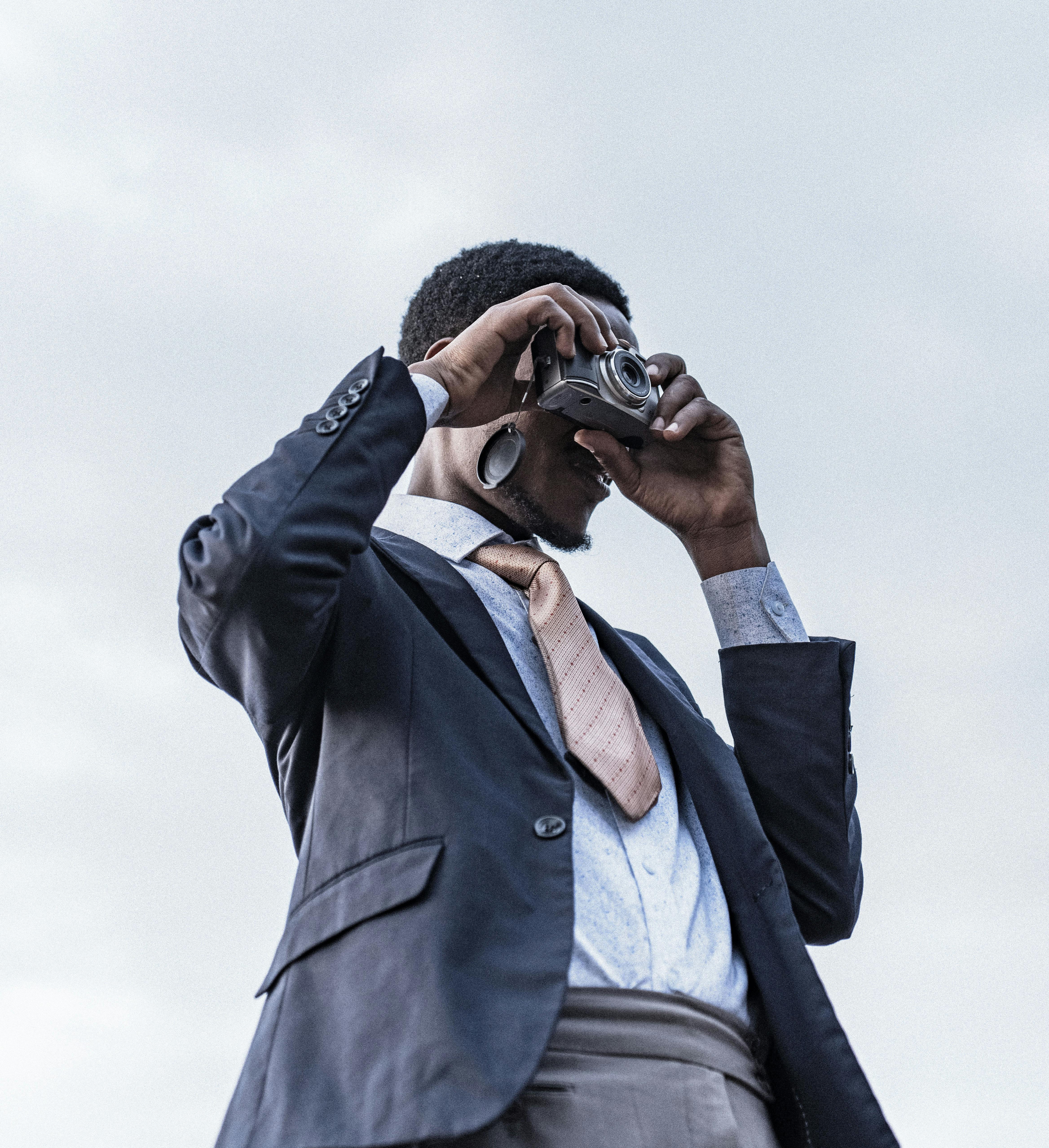 Man dressed in a suit using a vintage camera against a cloudy sky.