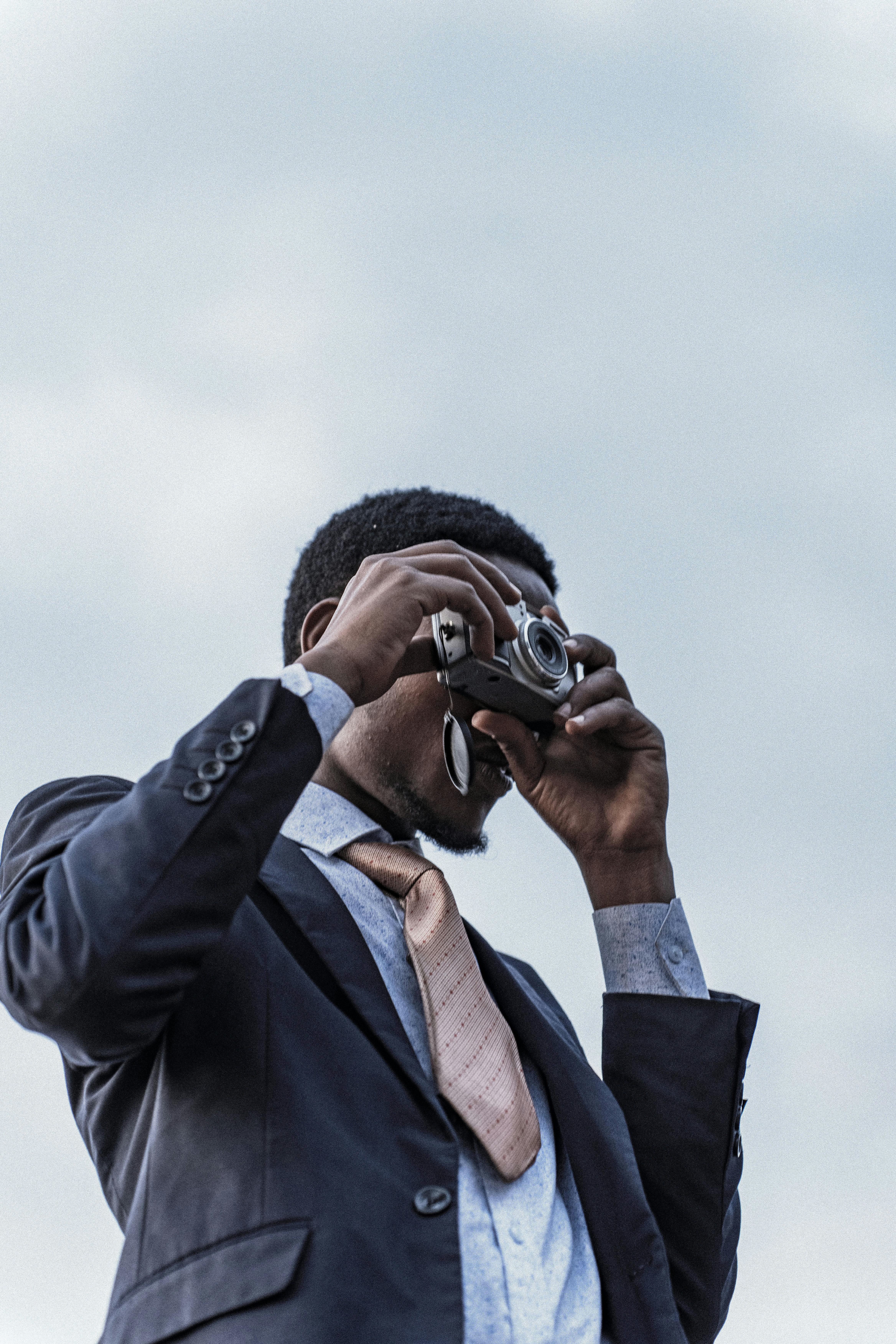 免费 一位身着西装、衣着考究的男士正在使用一台老式相机对着晴朗的天空拍照。 素材图片