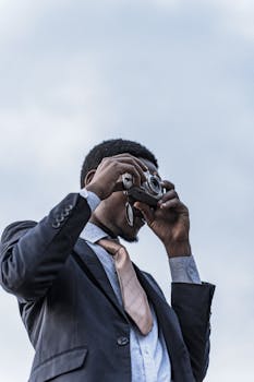 A well-dressed man in a suit uses a vintage camera against a clear sky.