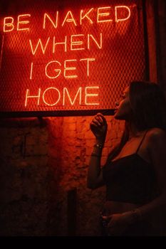 Fashionable woman standing by neon sign in a dimly lit urban environment.
