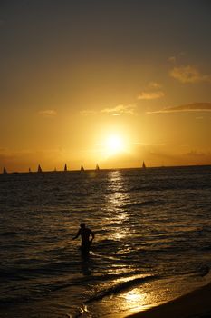 A captivating sunset view over the ocean with a silhouette of a person wading in the water, sailing boats on the horizon.