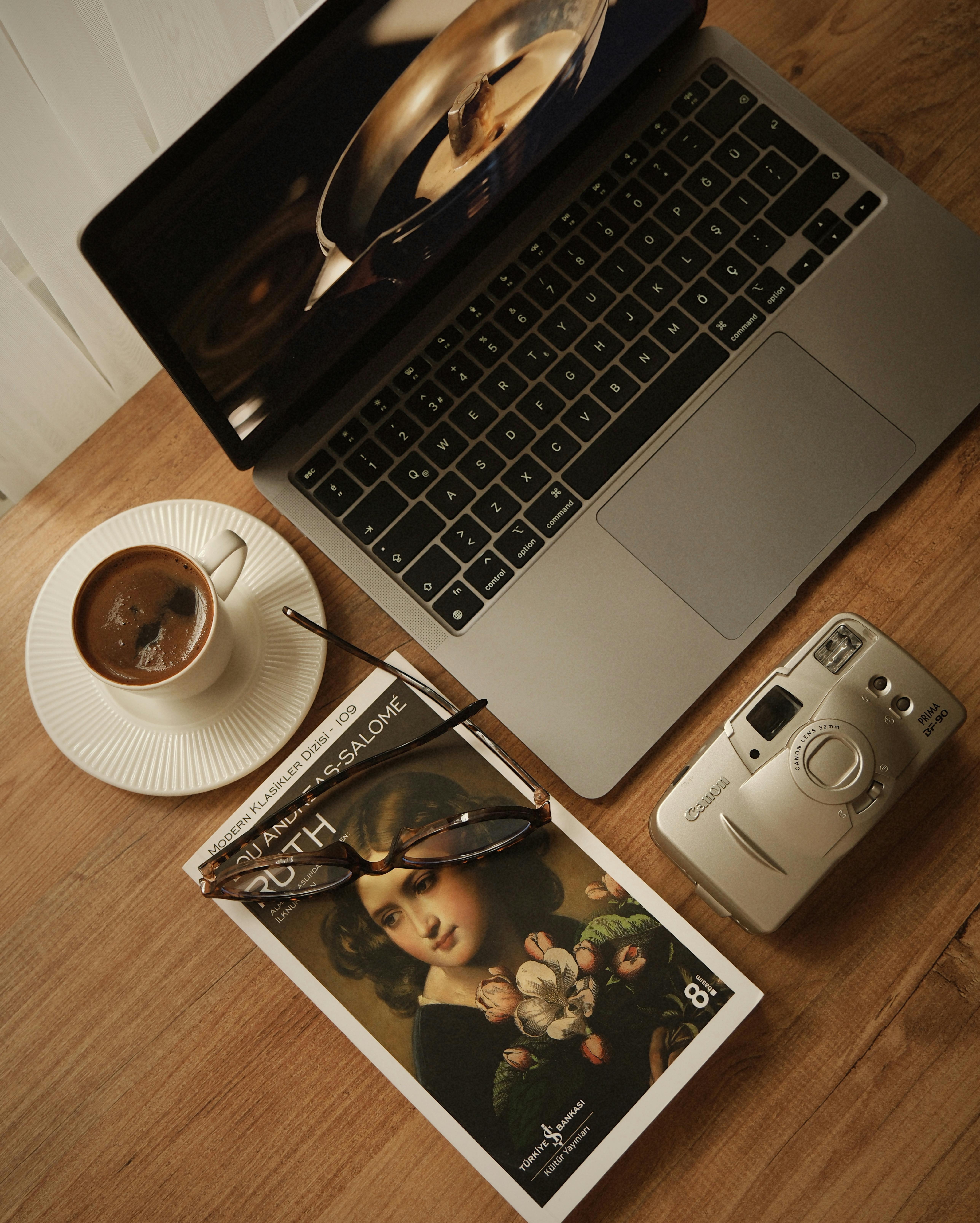 Gratis Un comodo tavolino con caffè, una macchina fotografica d'epoca, un libro aperto e un computer portatile su una scrivania di legno. Foto a disposizione