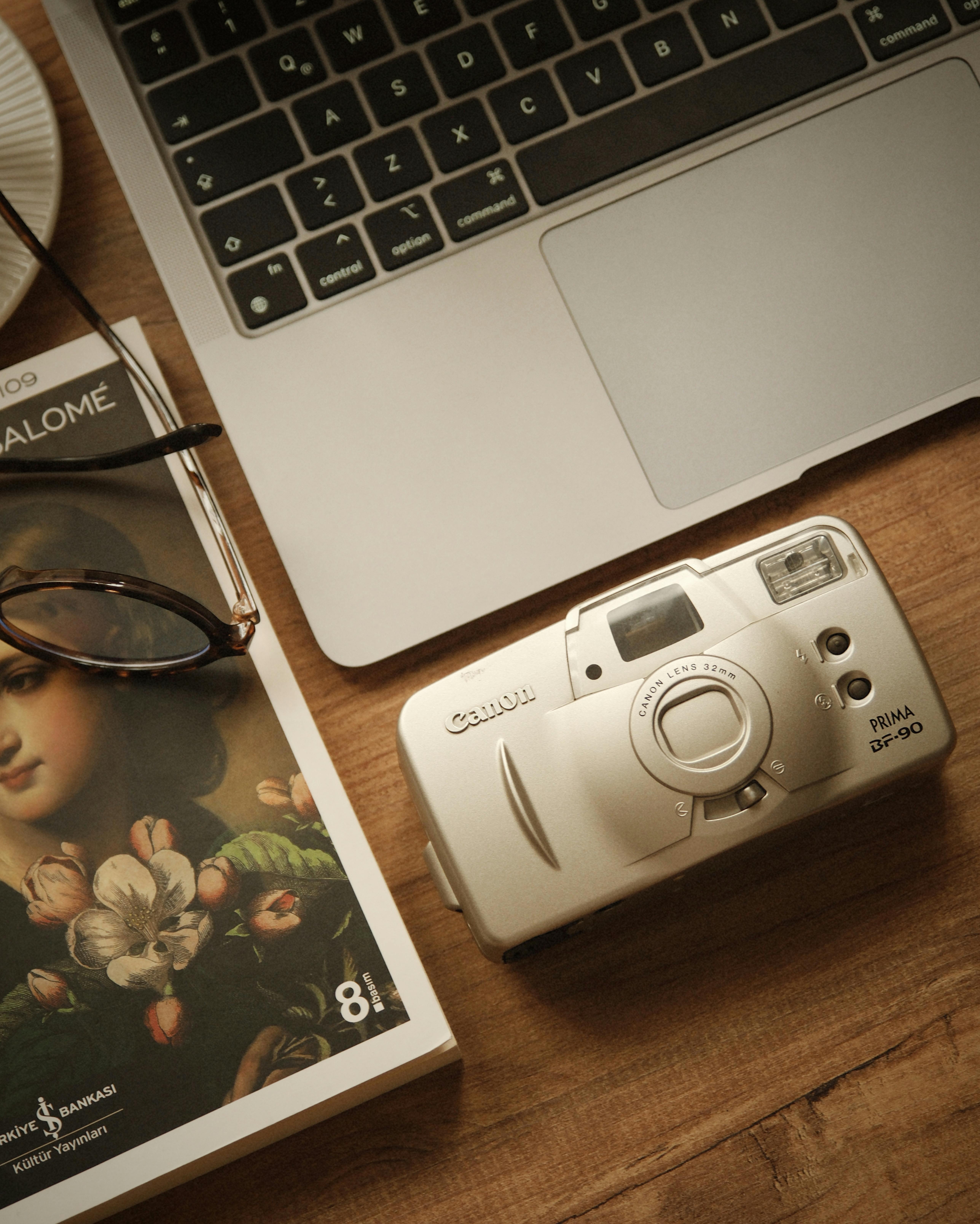 Miễn phí Hình ảnh nhìn từ trên xuống của một chiếc máy ảnh phim cổ điển, một cuốn sách và một chiếc máy tính xách tay trên một chiếc bàn gỗ, gợi lên cảm giác hoài cổ. Ảnh lưu trữ