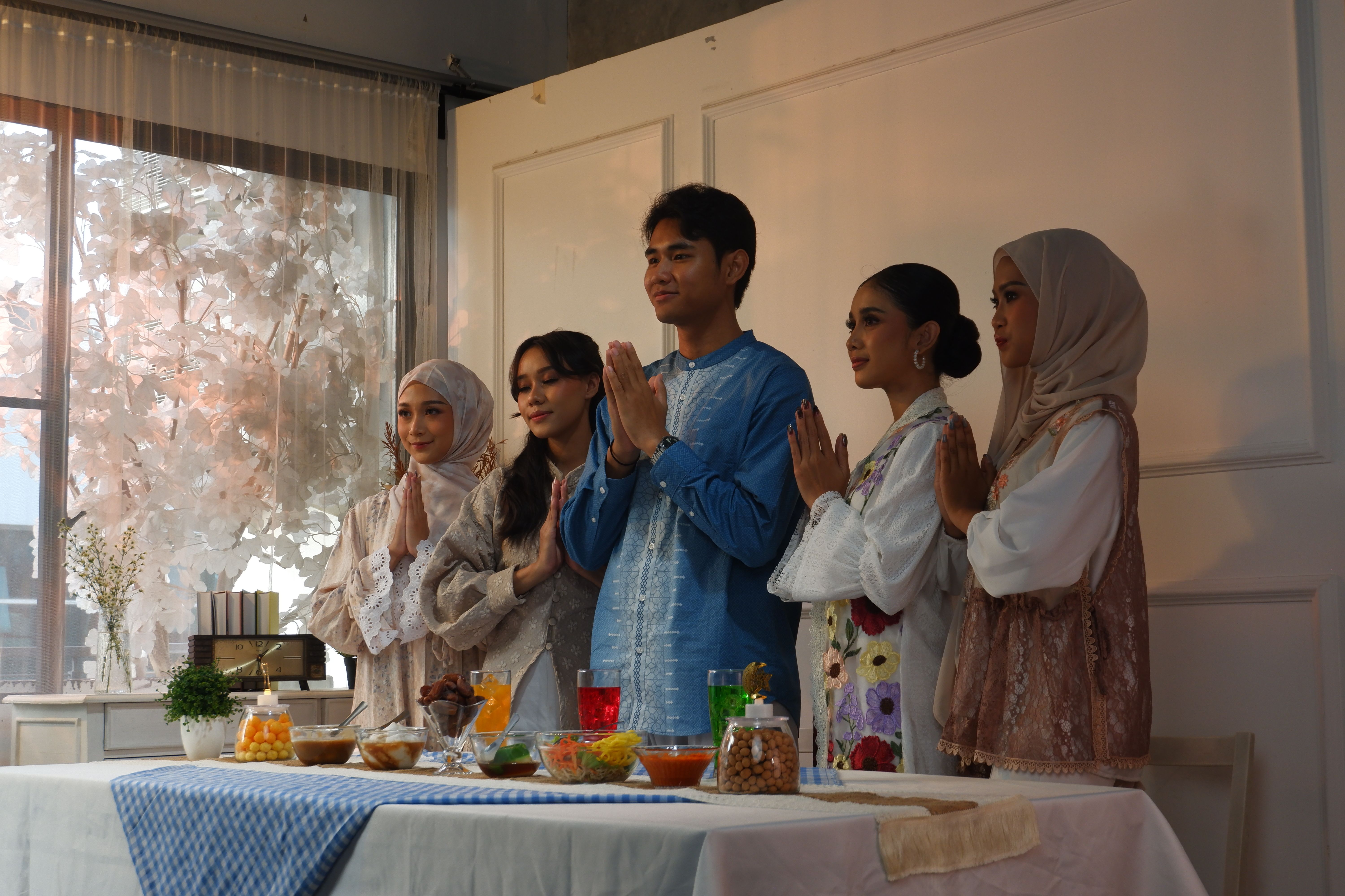 Diverse group in traditional attire praying at a festive dining table indoors, daylight setting.