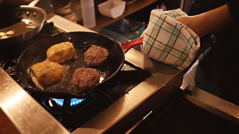 Close-up of two burgers and two patties cooking in a pan on a stove, kitchen scene.