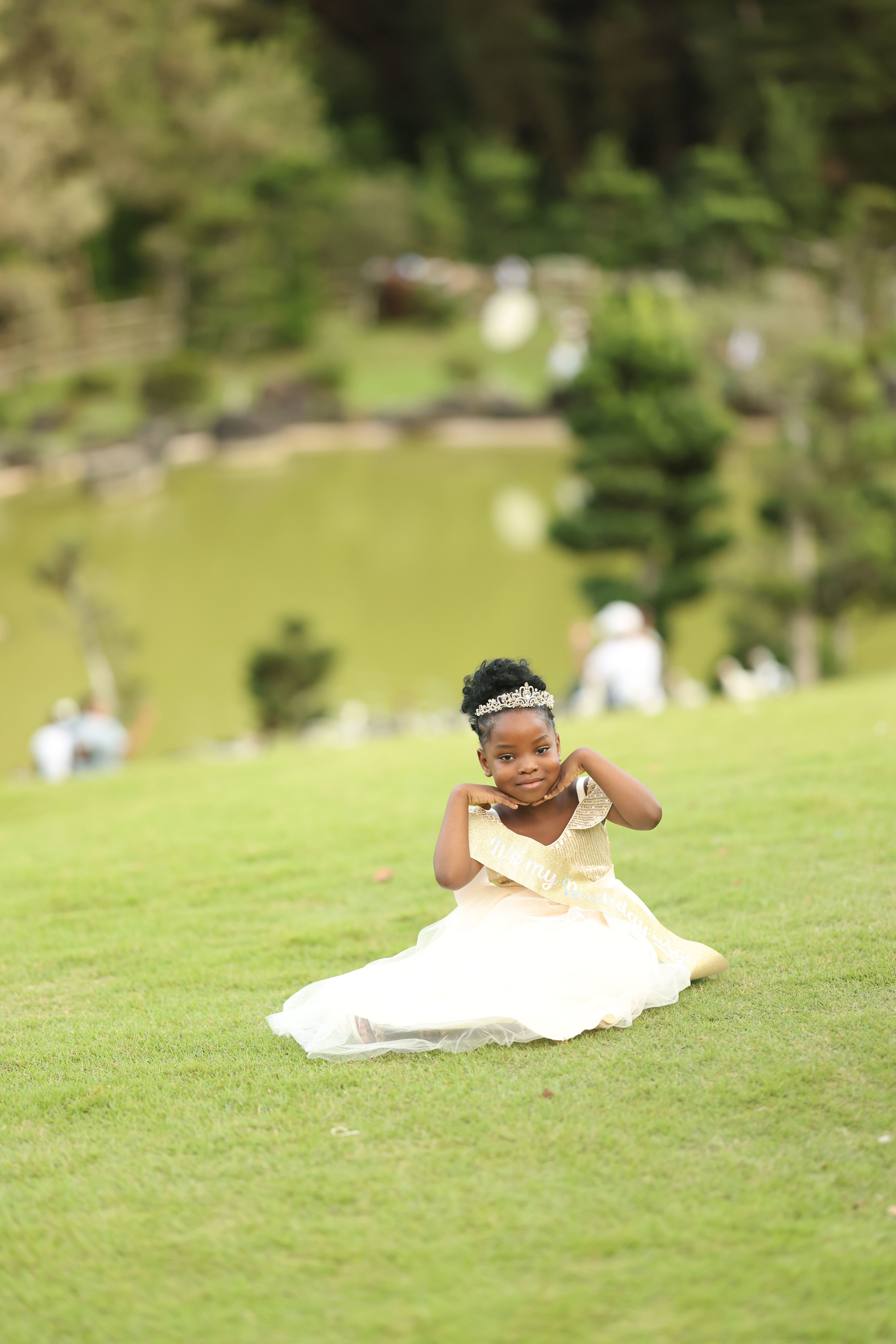 A young girl in a light dress posing outdoors on lush green grass by a pond.