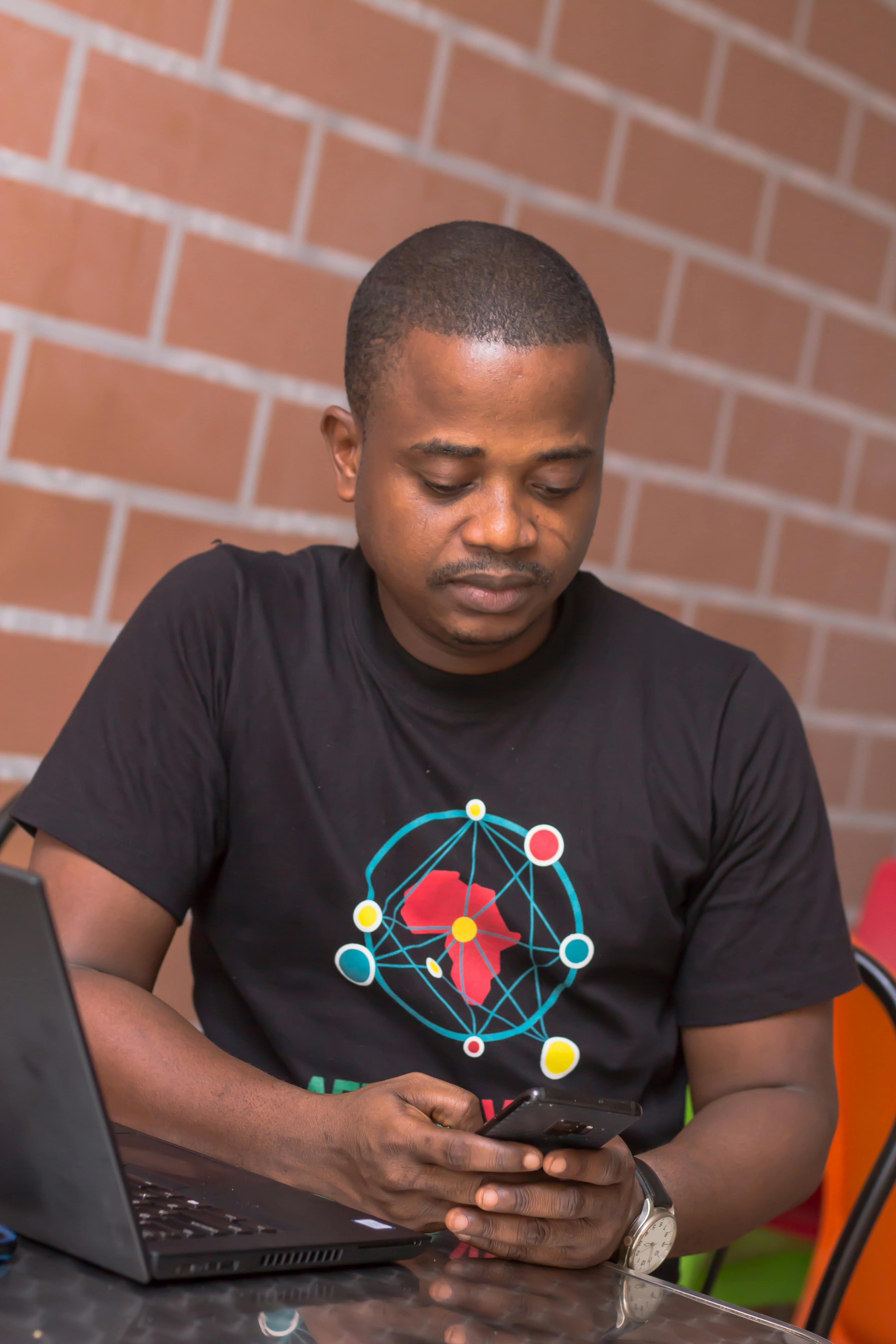 一名身穿黑色衬衫的男子坐在桌旁，一手拿着笔记本电脑，一手使用着智能手机