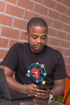 一名身穿黑色衬衫的男子坐在桌旁，一手拿着笔记本电脑，一手使用着智能手机