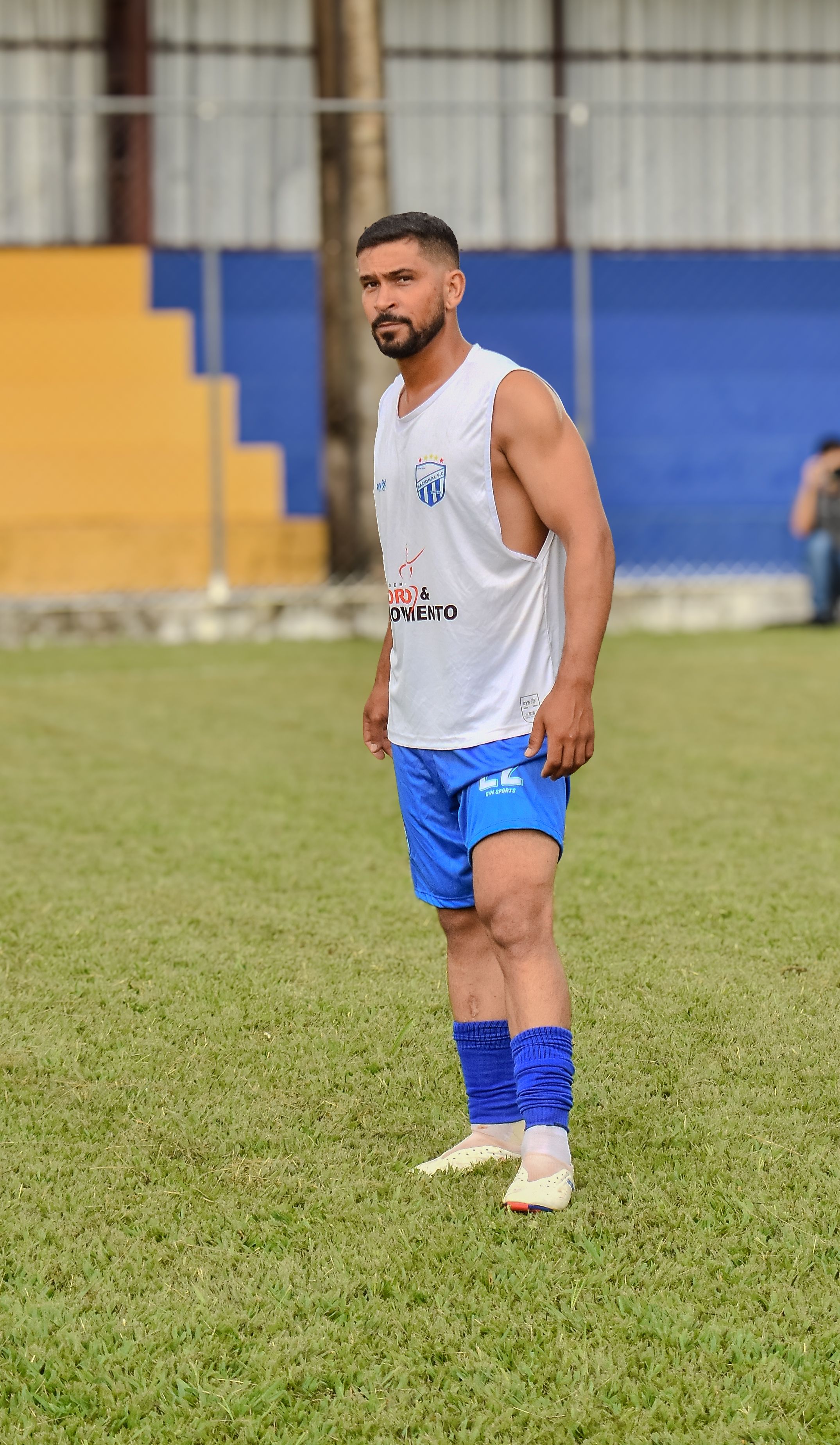 An adult male soccer player stands on a grassy field, wearing a blue and white uniform.