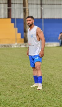An adult male soccer player stands on a grassy field, wearing a blue and white uniform.