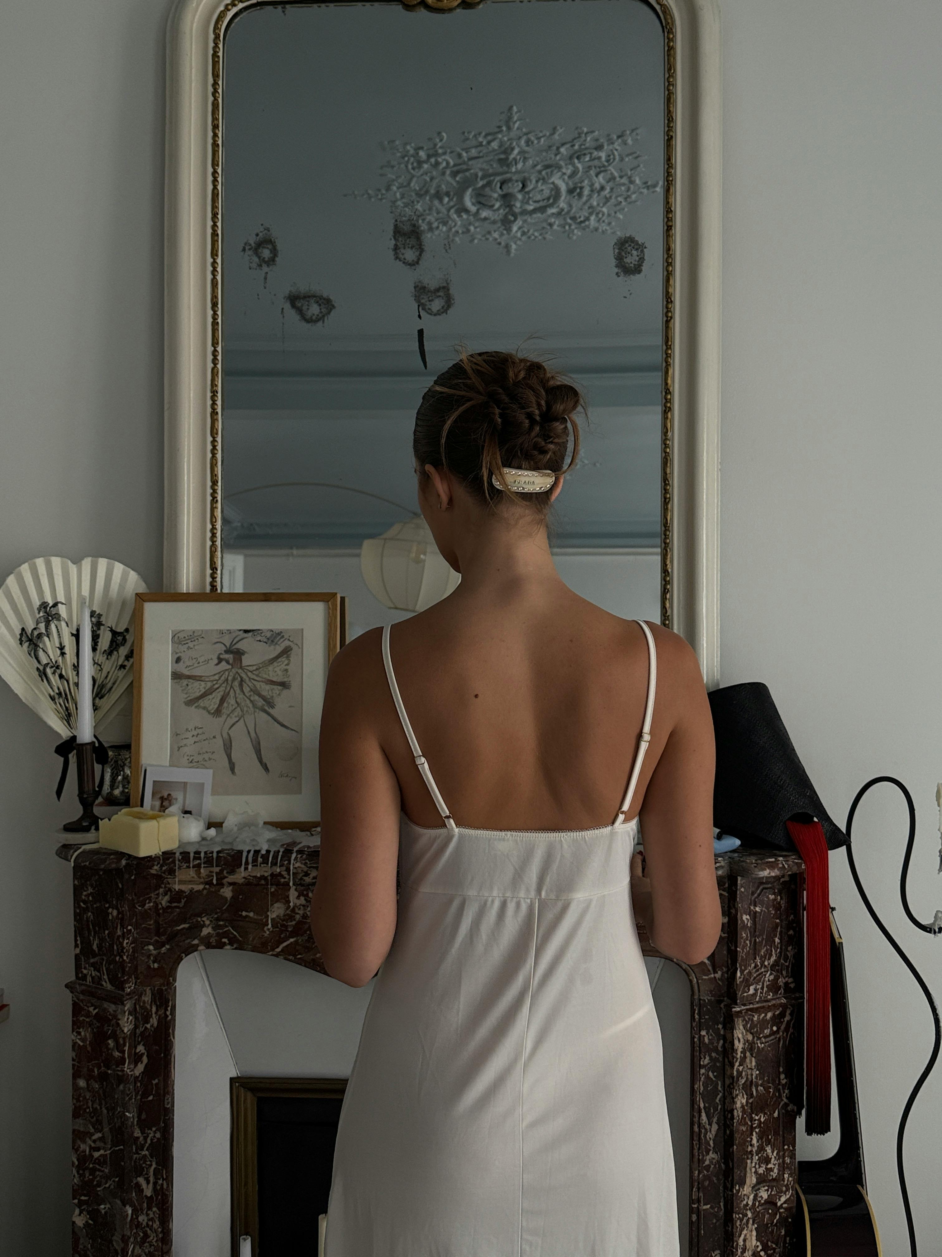 Free A woman in an elegant white dress stands in front of a mirror indoors, capturing a serene and timeless moment. Stock Photo