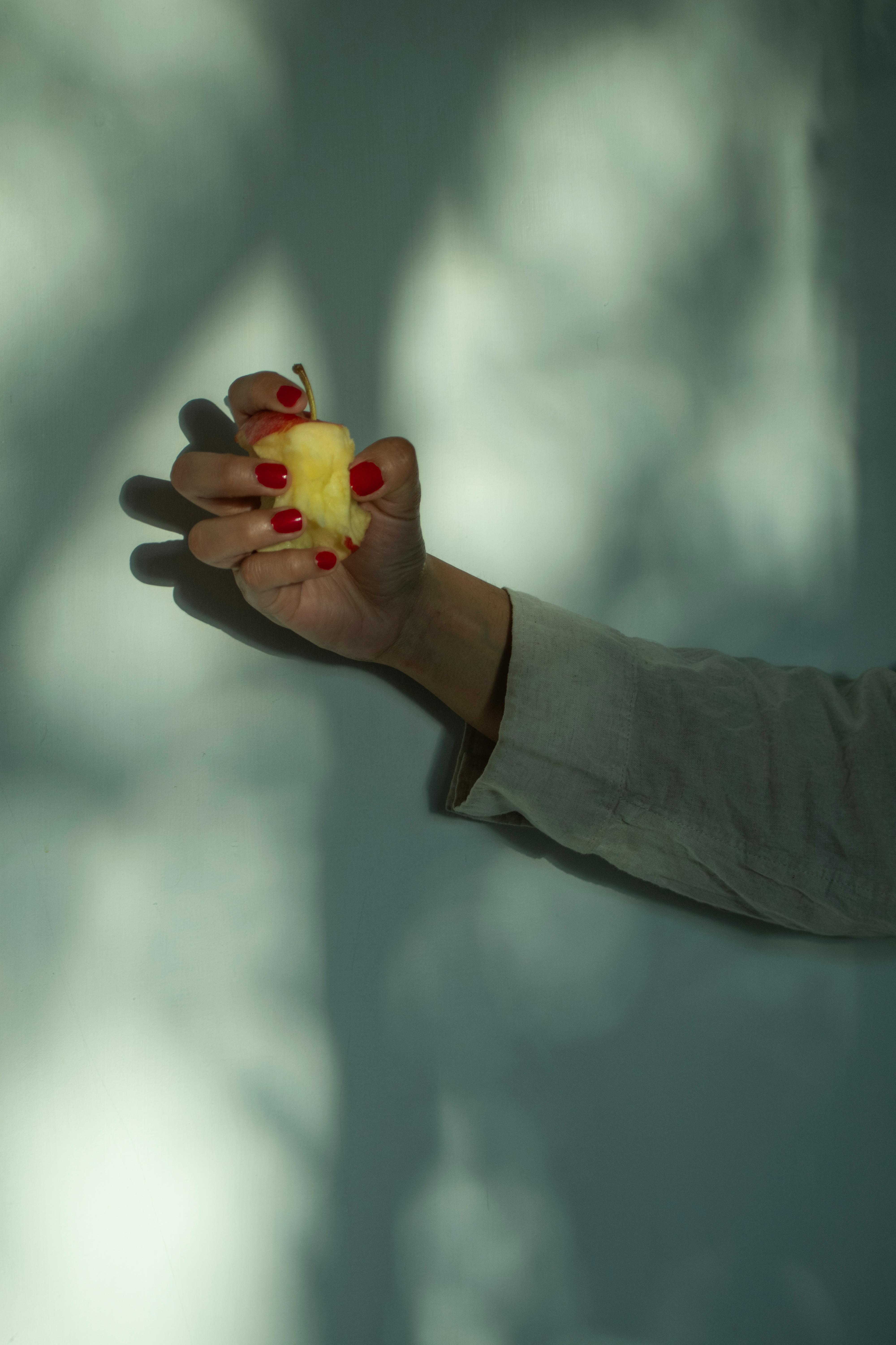 gratis Een hand met rode nagels houdt een geschilde appel tegen een schaduwrijke muur, wat een dramatisch effect creëert. Stockfoto