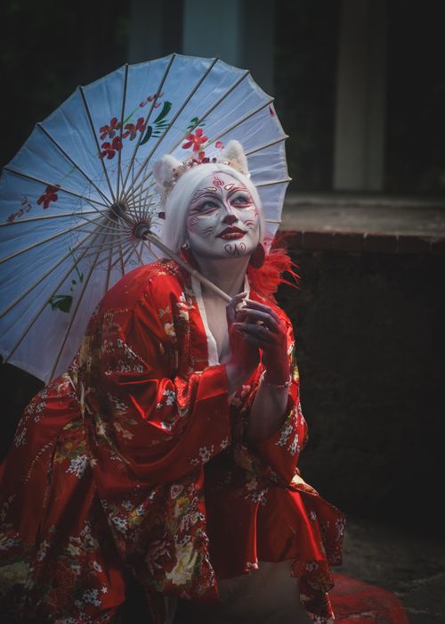 Free Artistic portrait of a person in geisha makeup wearing a red kimono and holding a parasol. Stock Photo