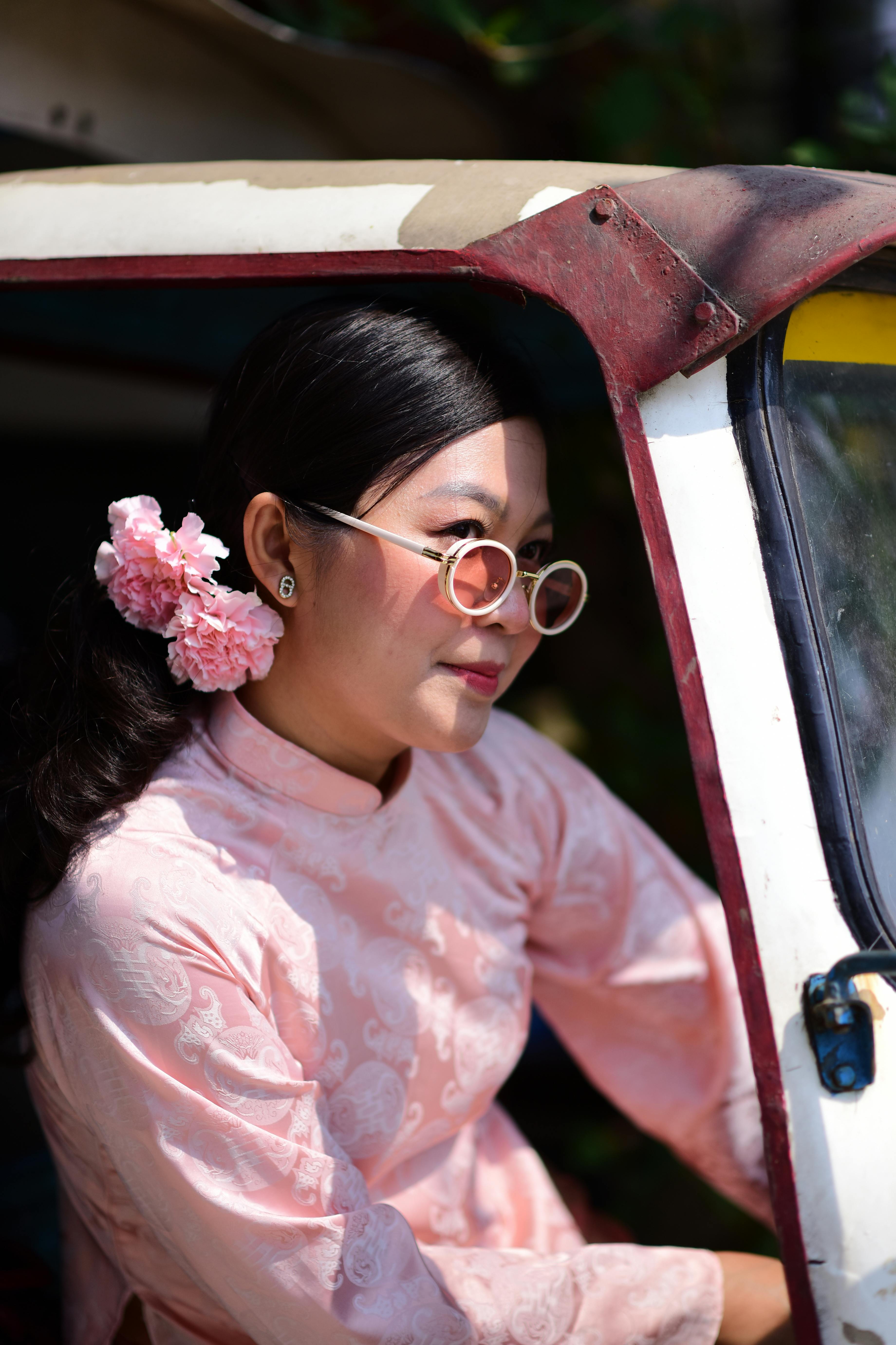 Zadarmo Žena v ružových šatách s kvetinovým vlasovým doplnkom sedí vo vozidle a má na sebe slnečné okuliare. Fotka z fotobanky