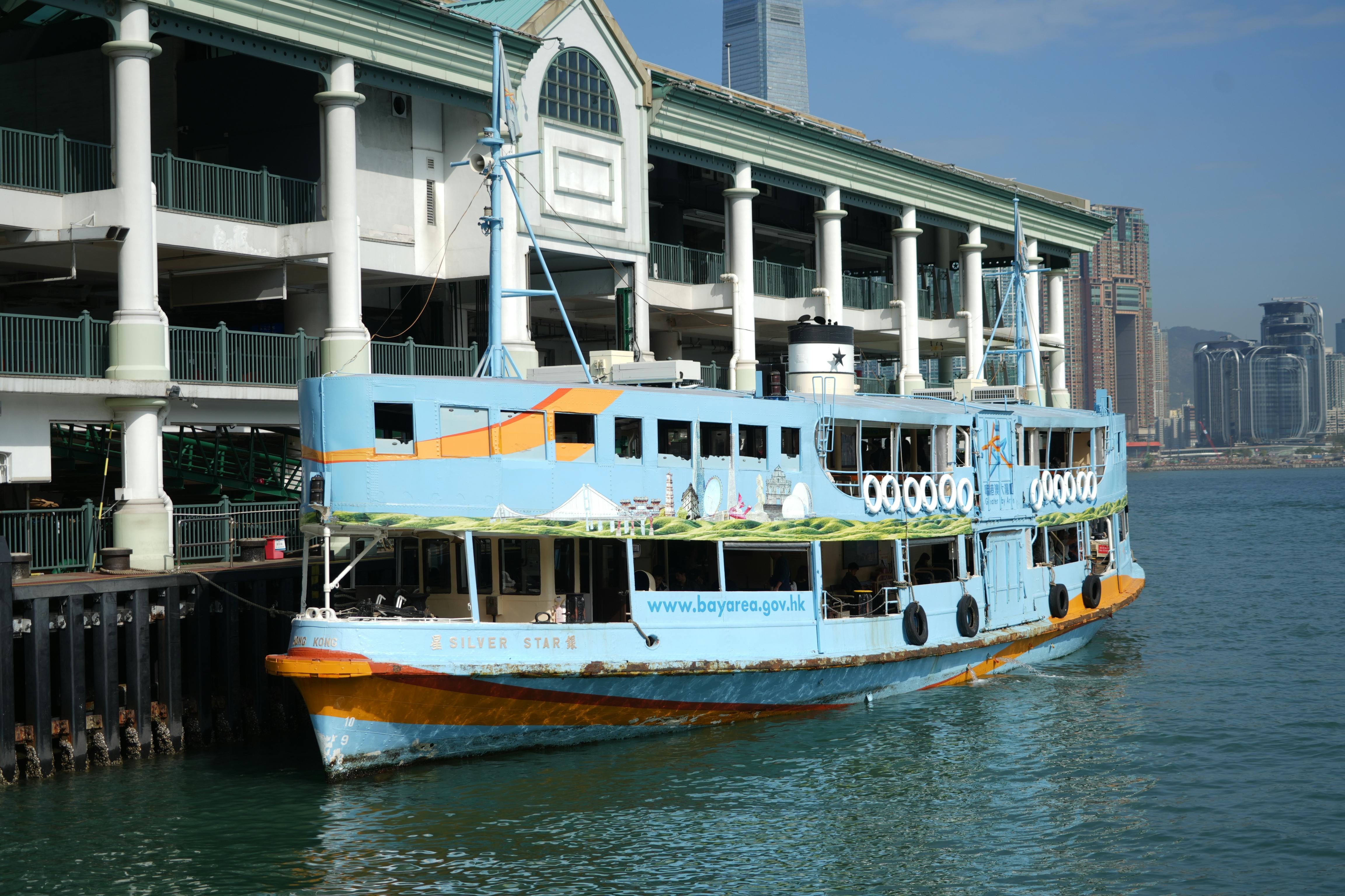A vibrant ferry boat docked at Central Pier on Hong Kong Island with city skyline in the background.