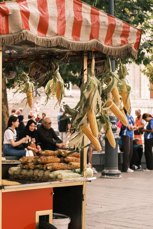 Free Street vendor selling roasted corn in a bustling city area with people relaxing nearby. Stock Photo