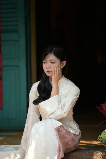 Young woman in white traditional Asian dress sitting contemplatively by ornate door.