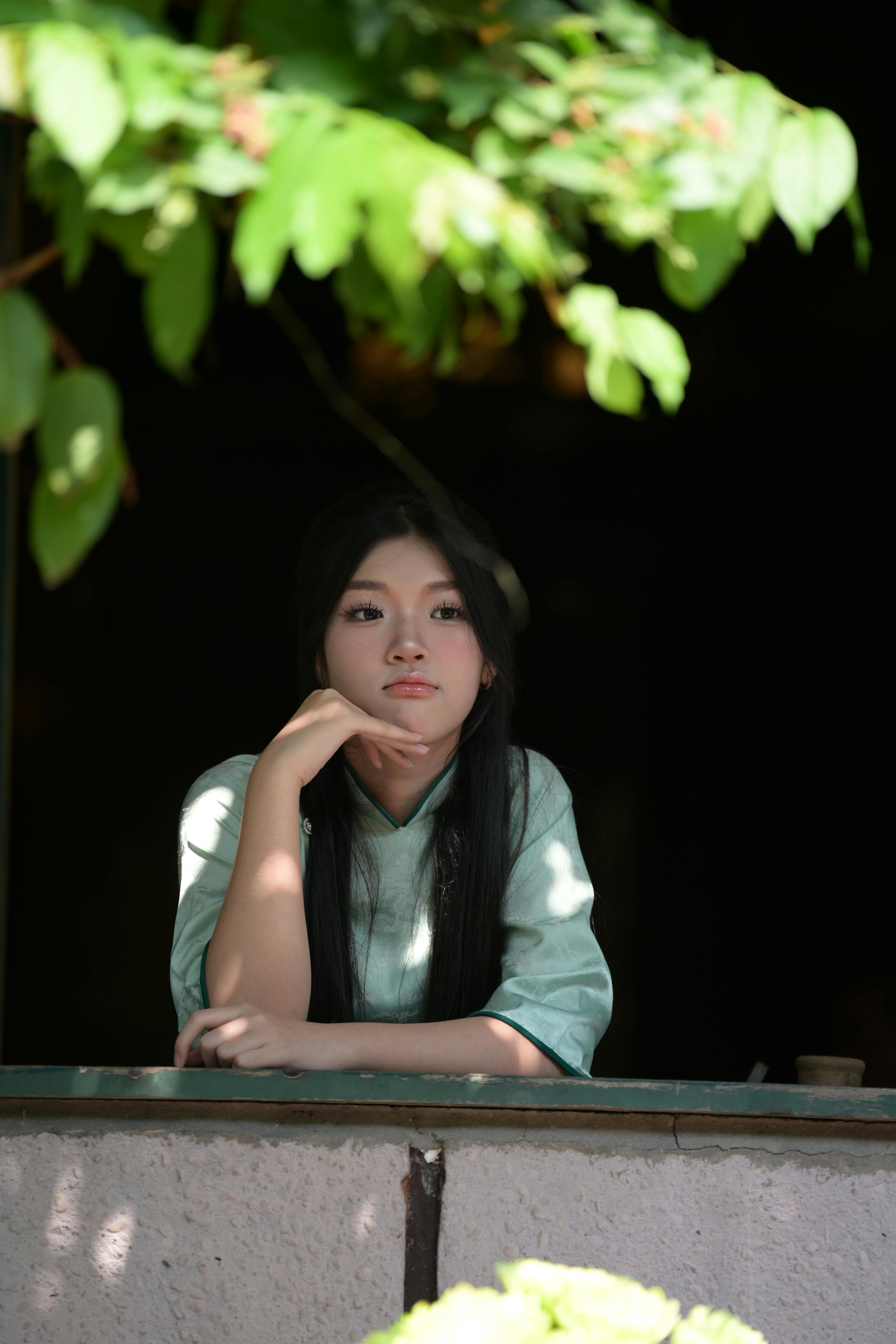 免费 一位身穿绿色连衣裙的年轻女子凝视着窗外，周围环绕着绿色植物。 素材图片