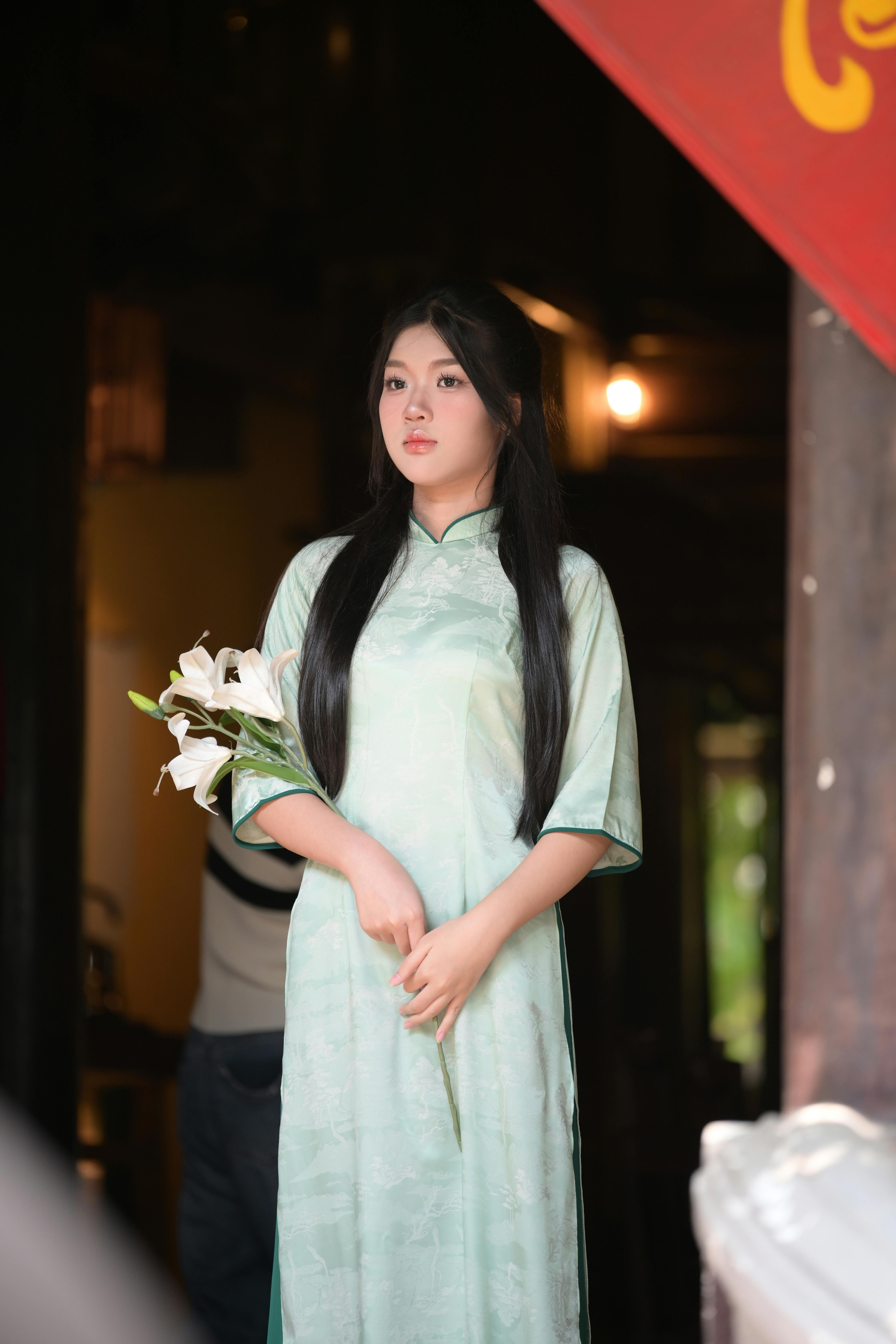 A woman in traditional attire holding lilies, standing indoors. The soft light creates a serene atmosphere.