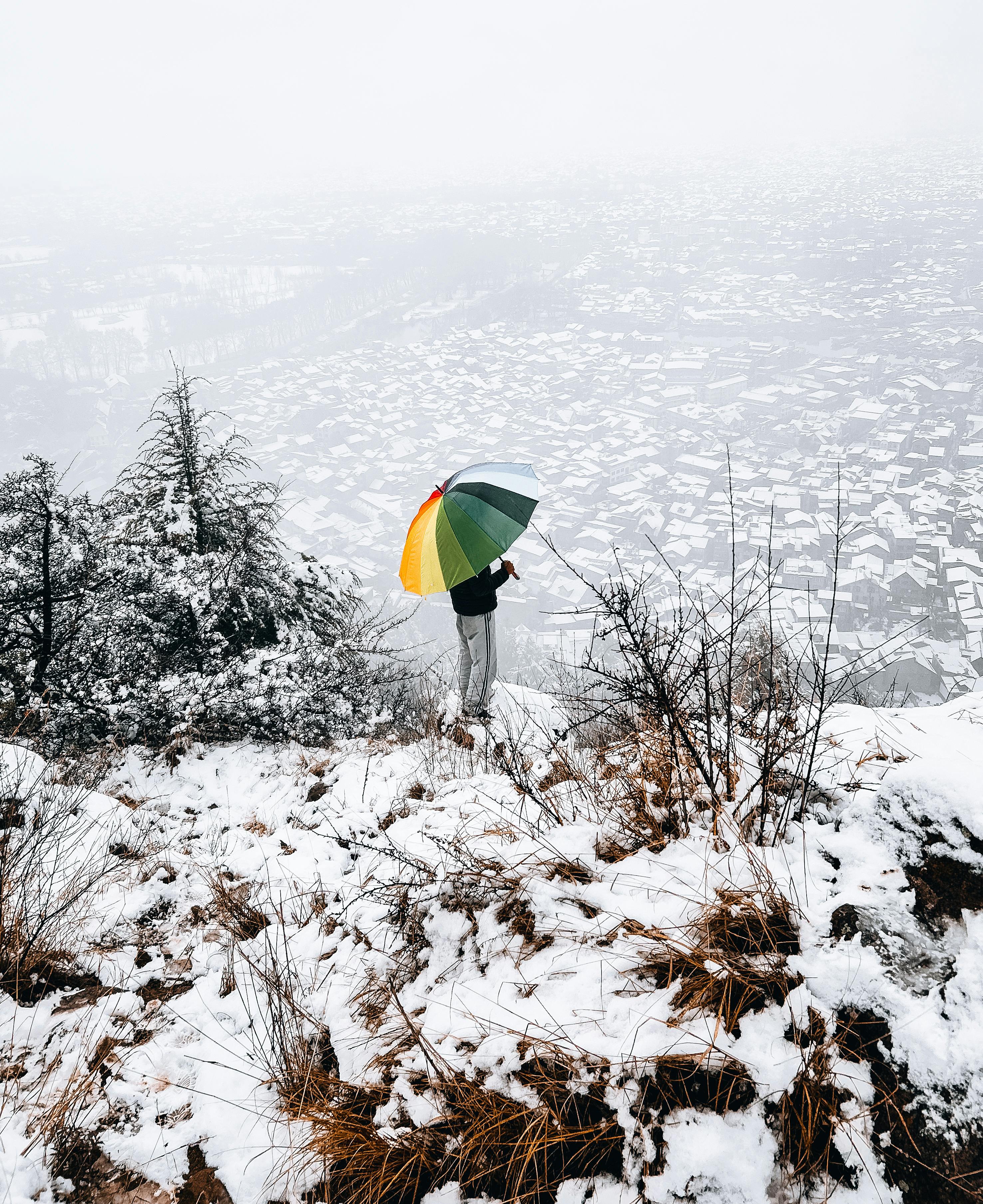 Person holding colorful umbrella standing on snowy hill with Srinagar city in the background.