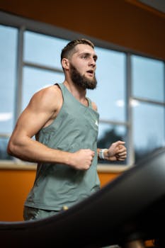 Focused young man jogging on a treadmill in a modern gym with large windows.