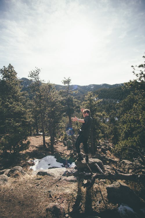 免費 穿黑夾克的男人站在白雲下綠樹附近的岩石地面上 圖庫相片
