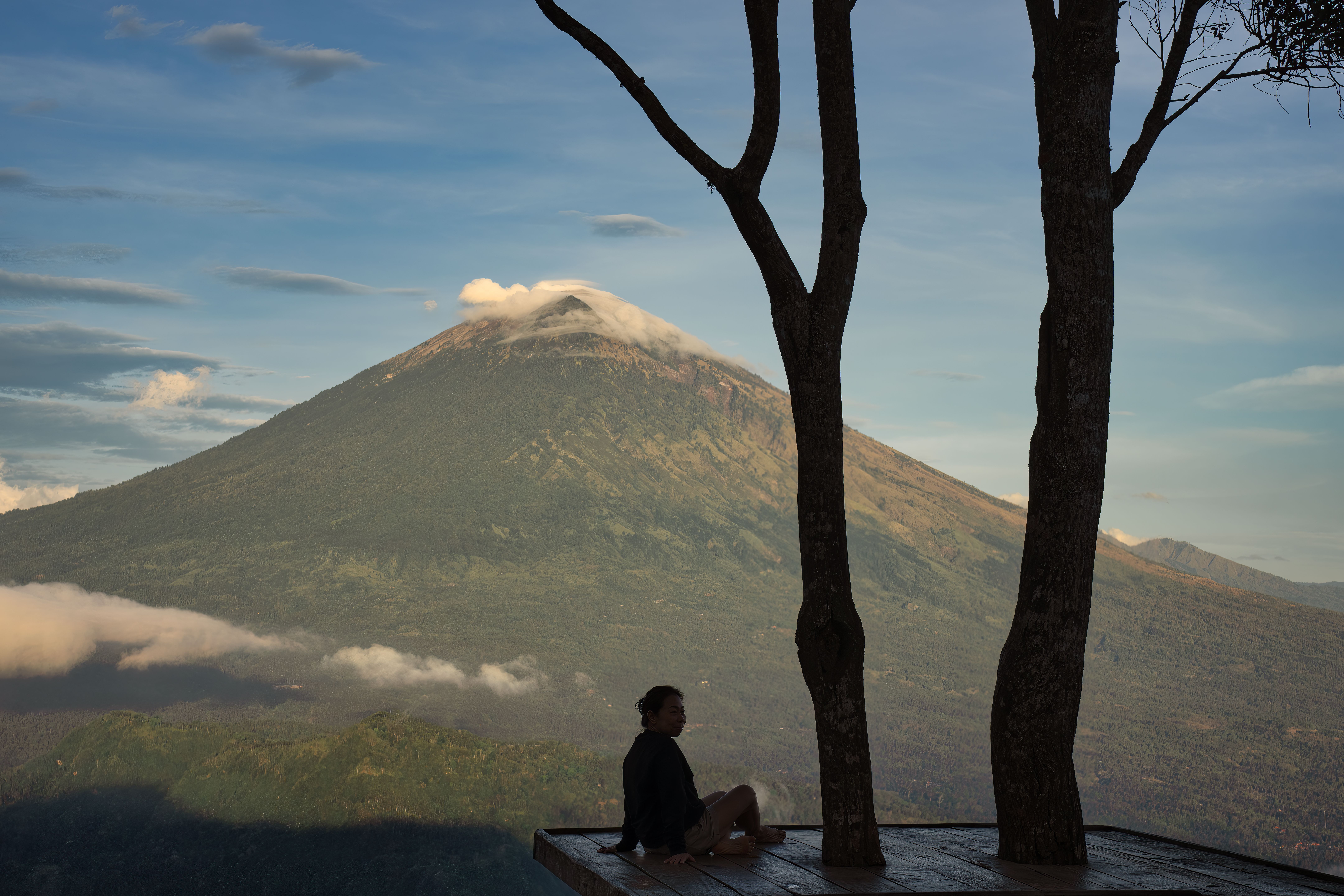 A serene scene with a person sitting near trees, overlooking Mount Agung in Bali.