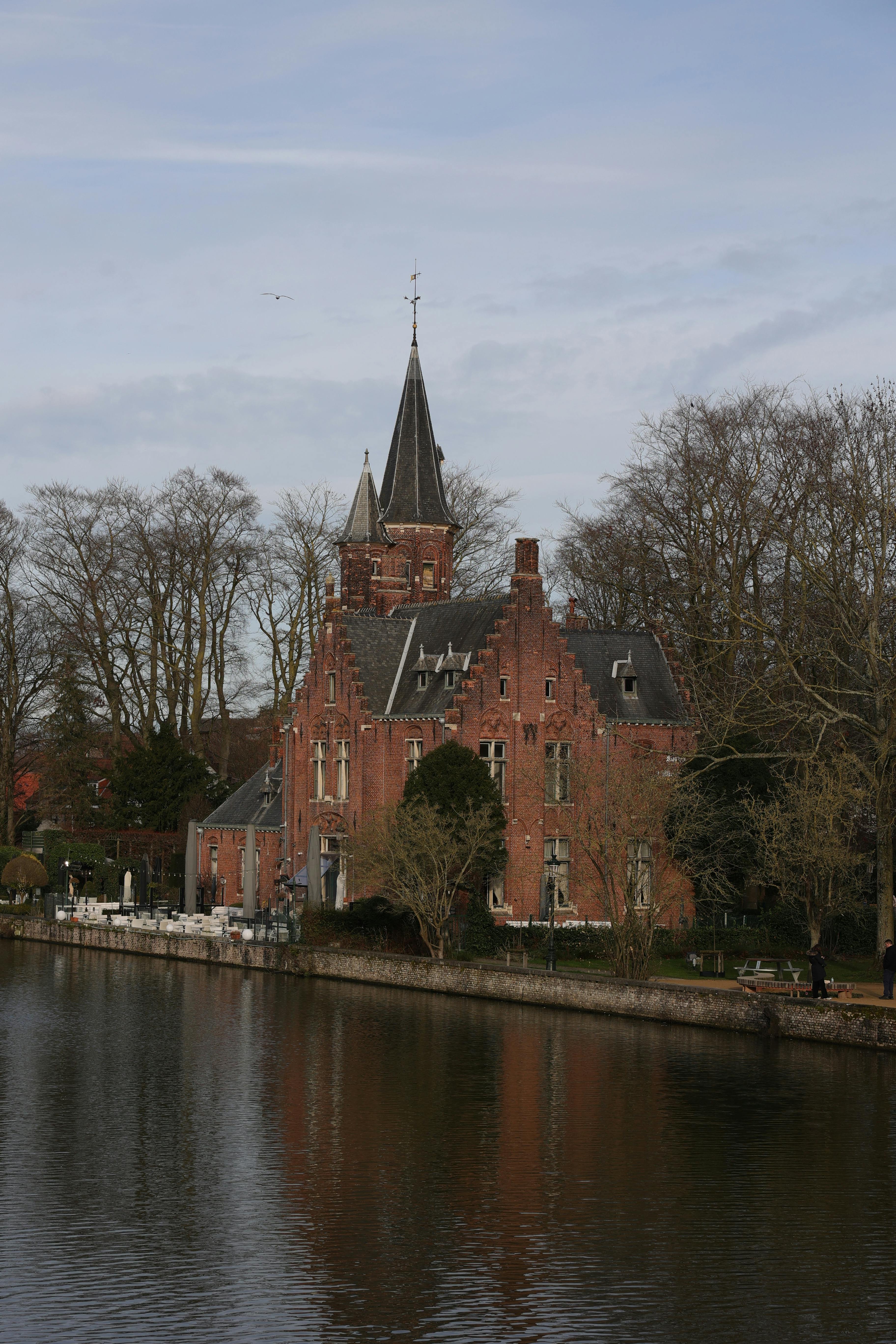 Gratis Bangunan bata yang menawan dengan pantulan menara di tepi kanal di Bruges, Belgia. Foto Stok