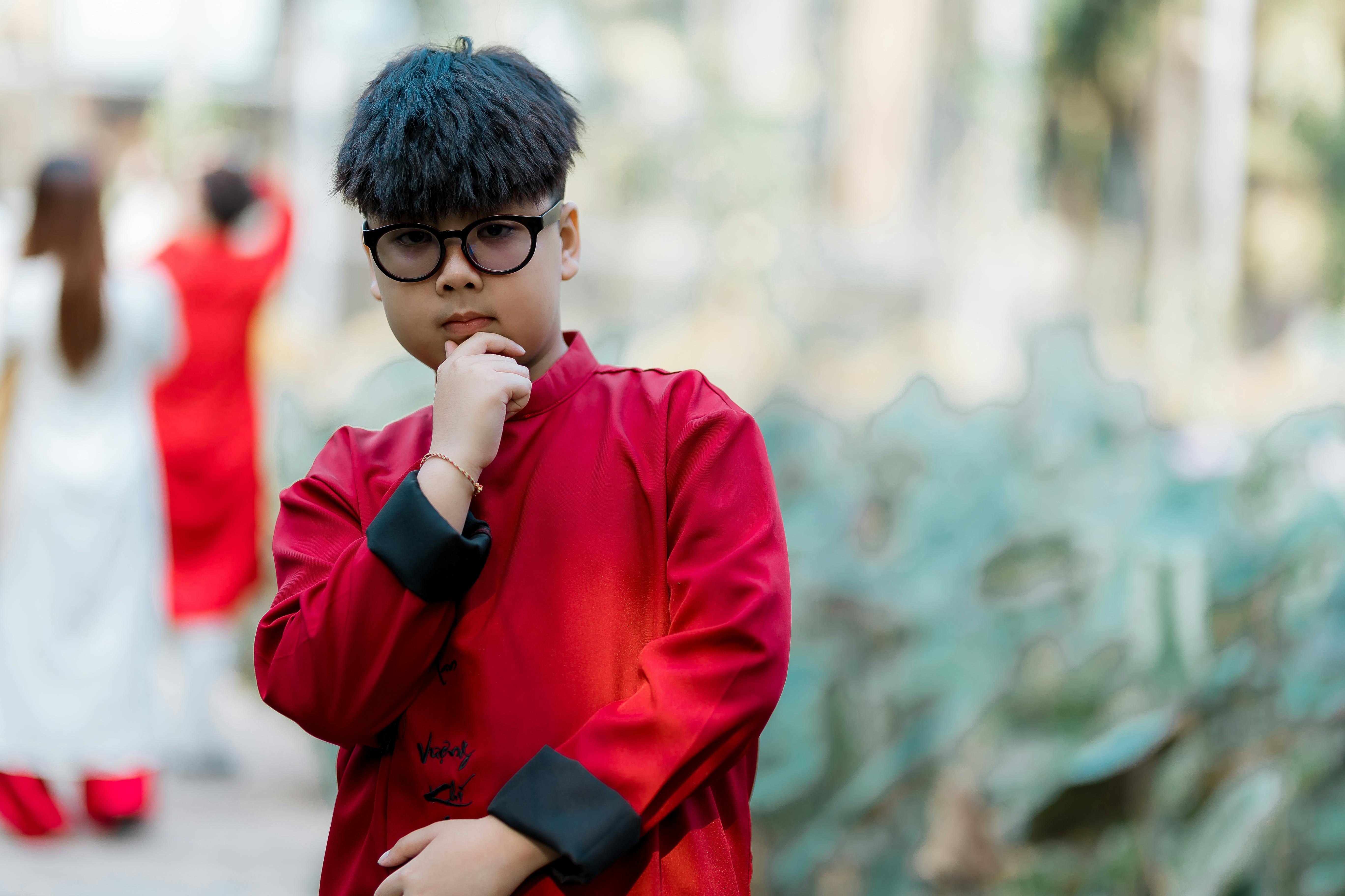 Bezpłatne Młody chłopiec w okularach i tradycyjnym czerwonym stroju zamyślony pozuje na zewnątrz. Zdjęcie z galerii