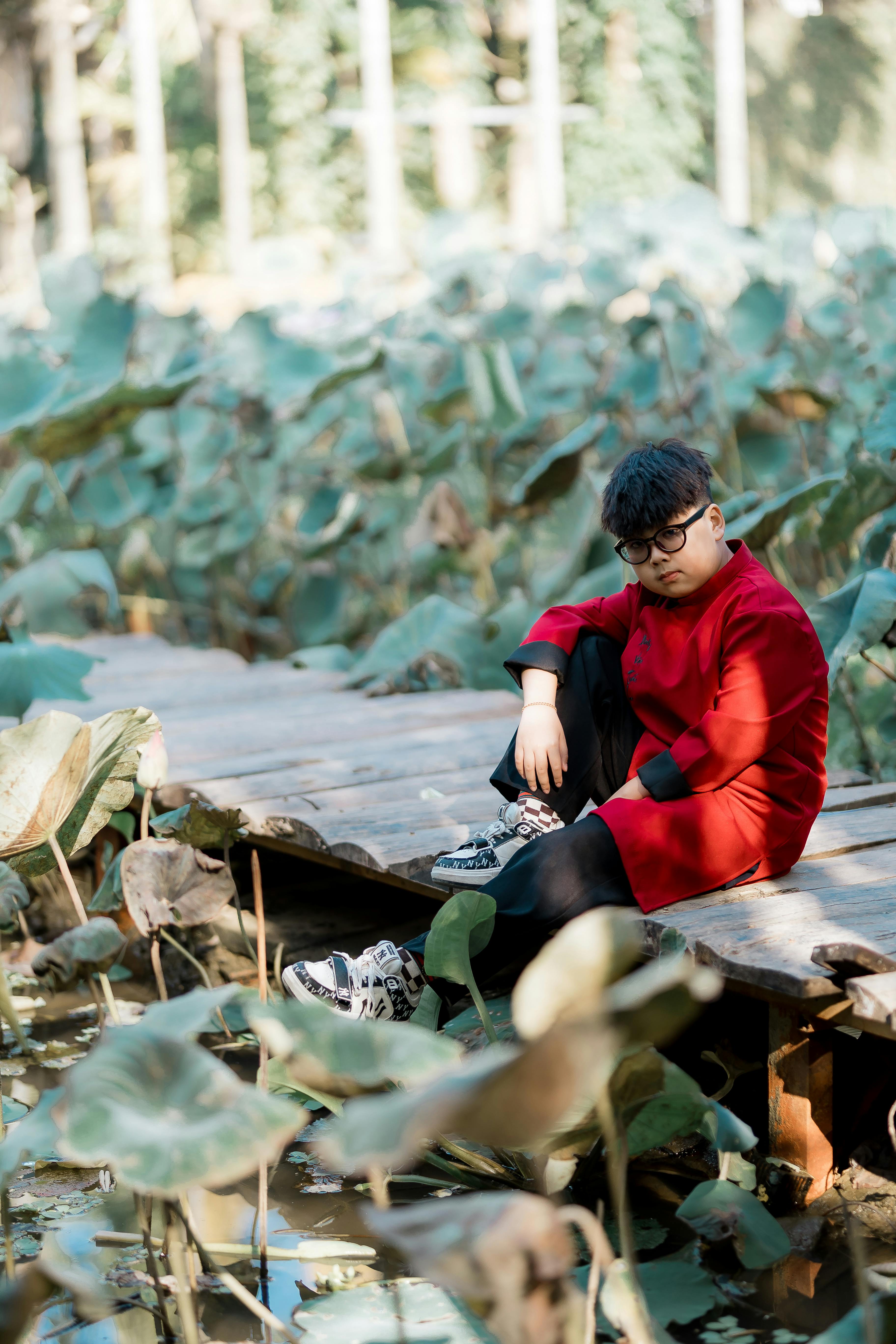 Безкоштовне Молодий чоловік у червоному пальто задумливо сидить на дерев'яній доріжці крізь пишну зелень. стокове фото