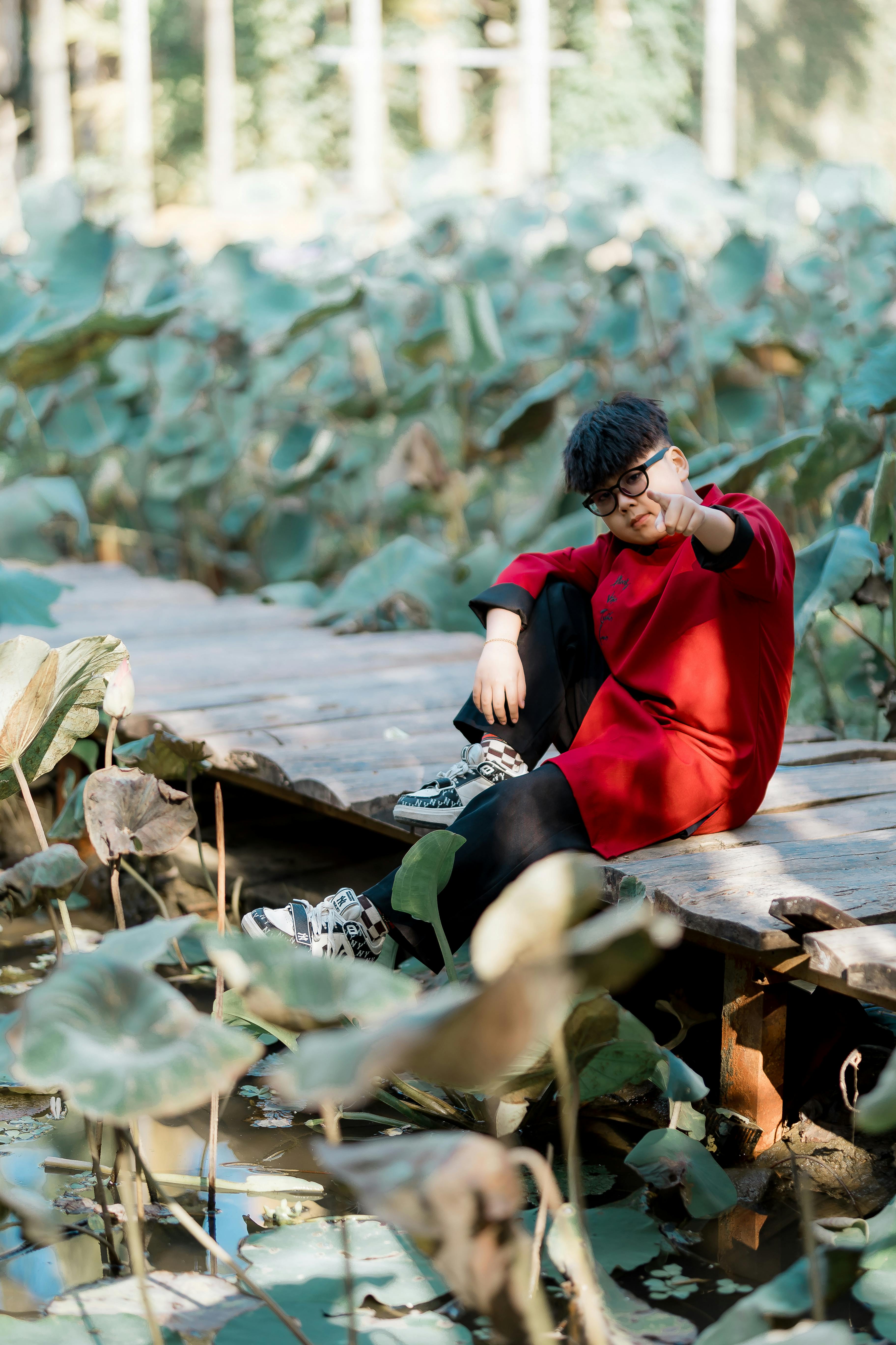 Gratis Adulto joven vestido de rojo sentado en un puente de madera en medio de exuberantes plantas verdes. Foto de stock