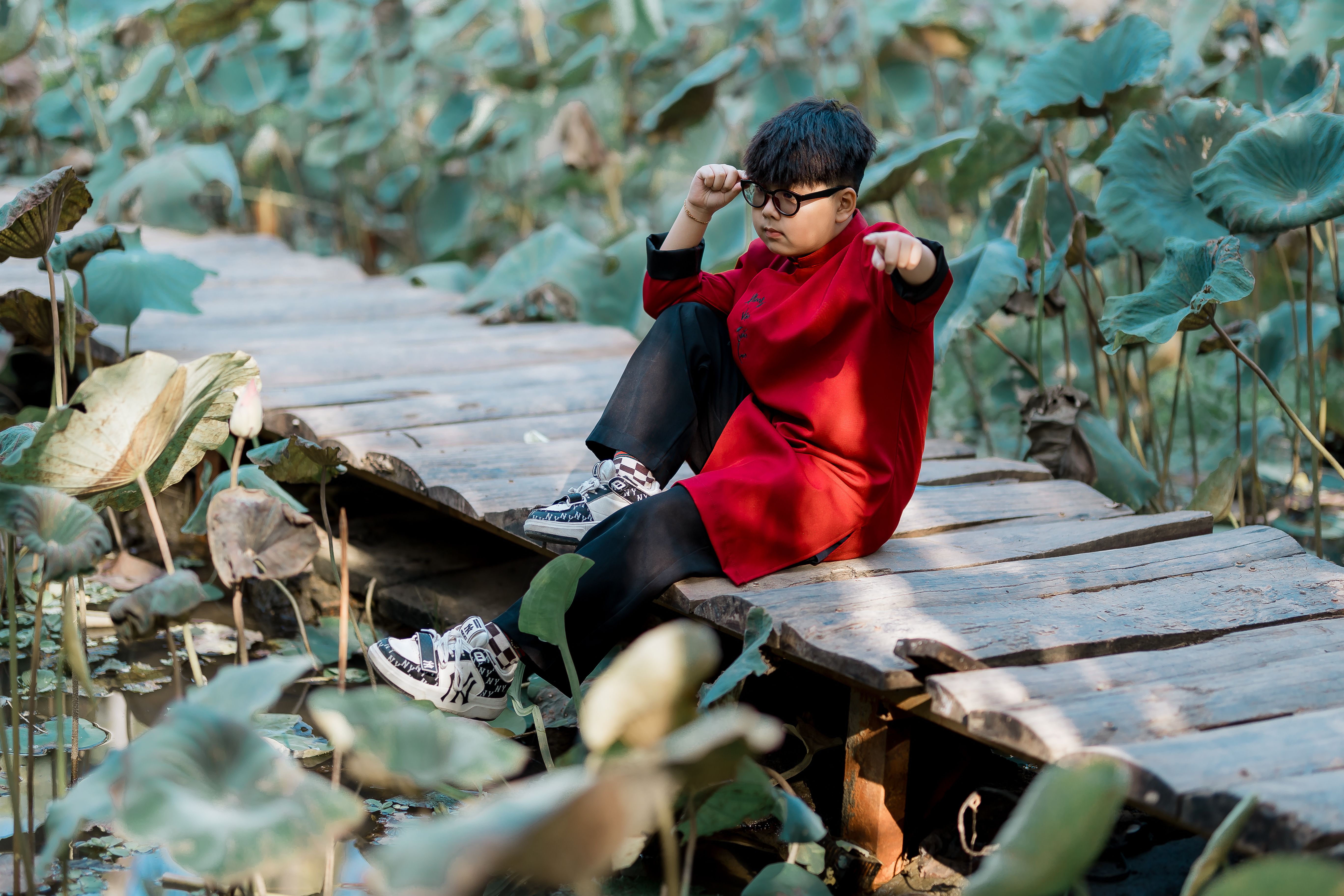 Young boy in red traditional attire poses on a wooden path amidst lush greenery.