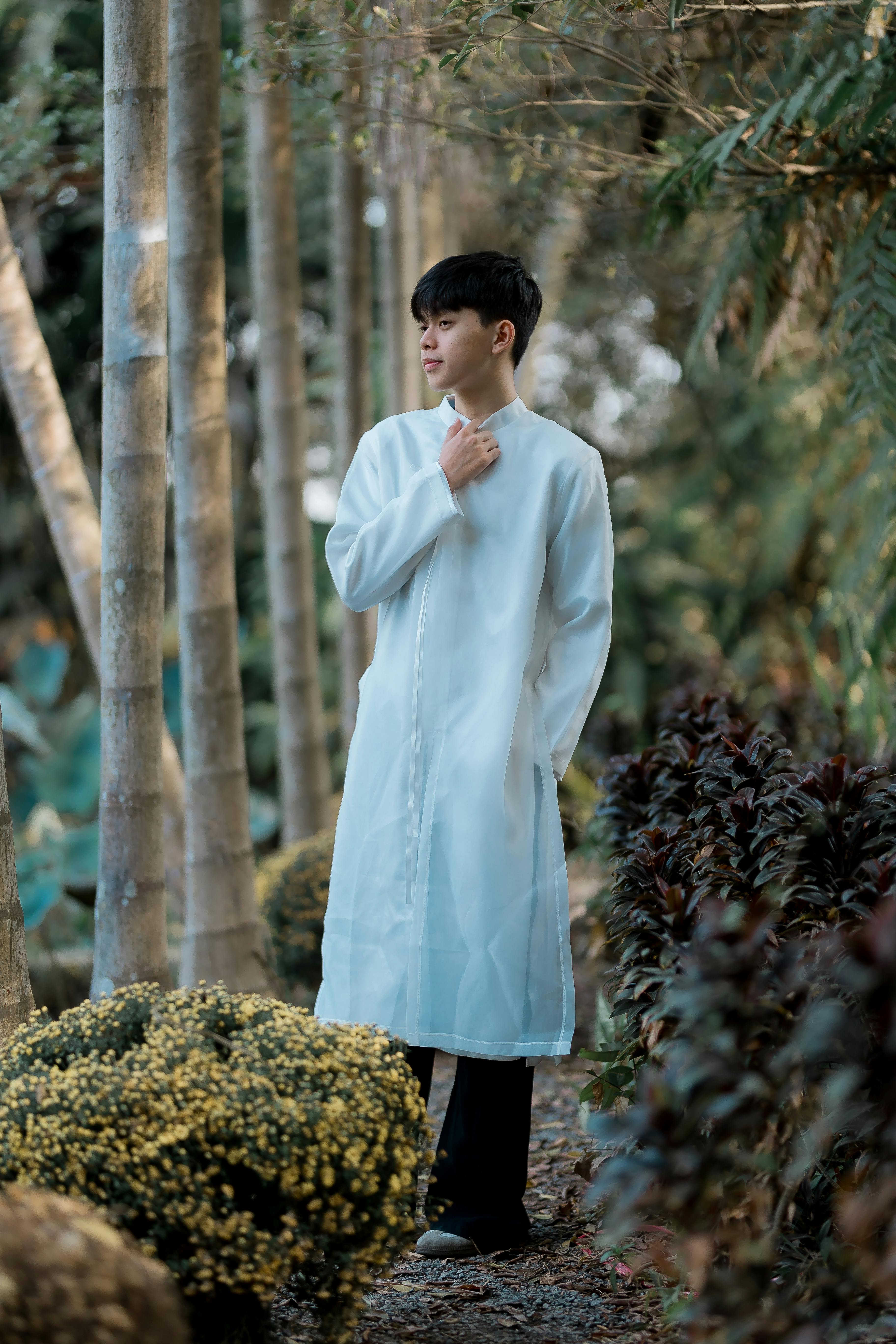 A young man in a white outfit stands on a tranquil garden pathway surrounded by lush vegetation.
