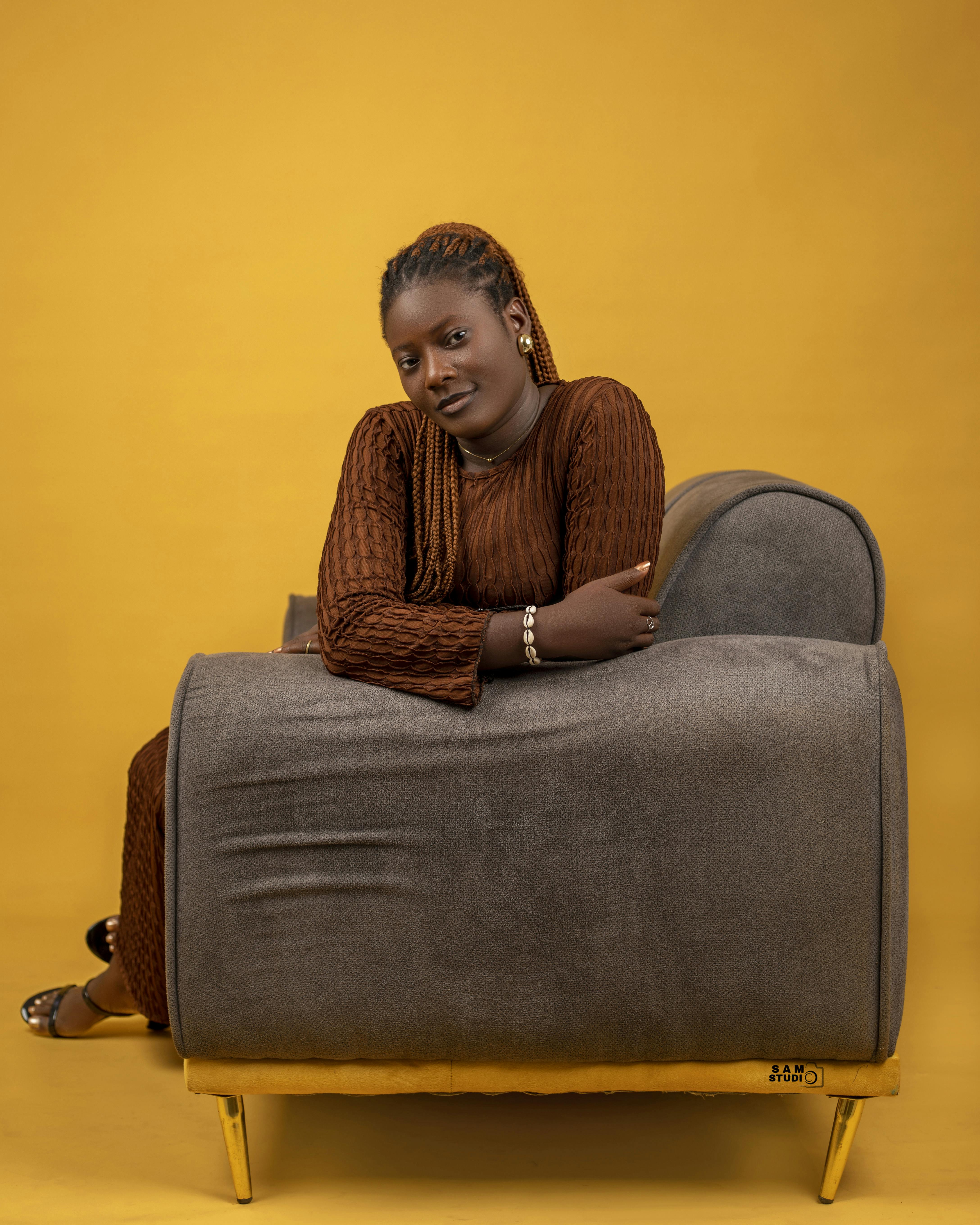 Gratis Elegante ritratto di una donna in abito marrone seduta su un divano con sfondo giallo. Foto a disposizione
