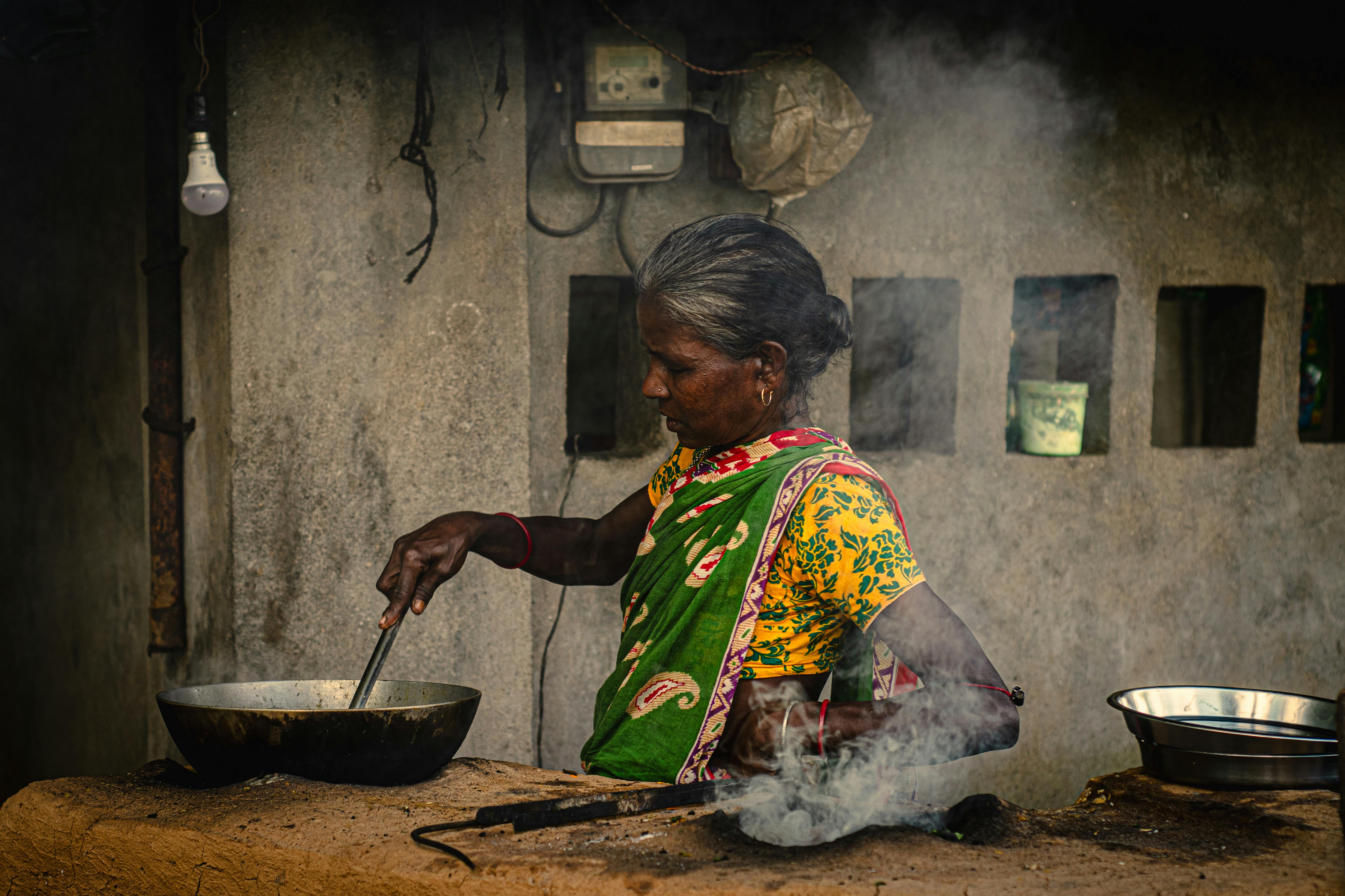 Gratis Seorang wanita pedesaan India dengan pakaian berwarna cerah menyiapkan makanan di luar ruangan menggunakan metode tradisional. Foto Stok