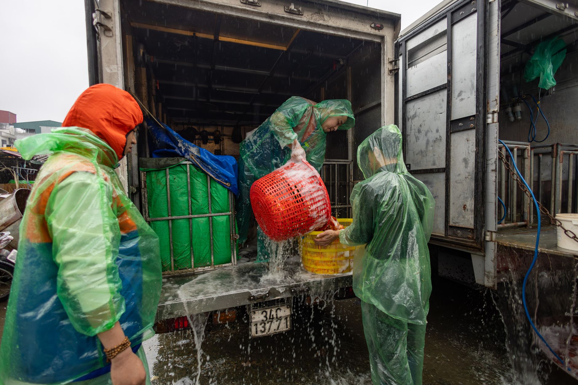Workers in raincoats unload goods in a rainy Vietnamese market scene.